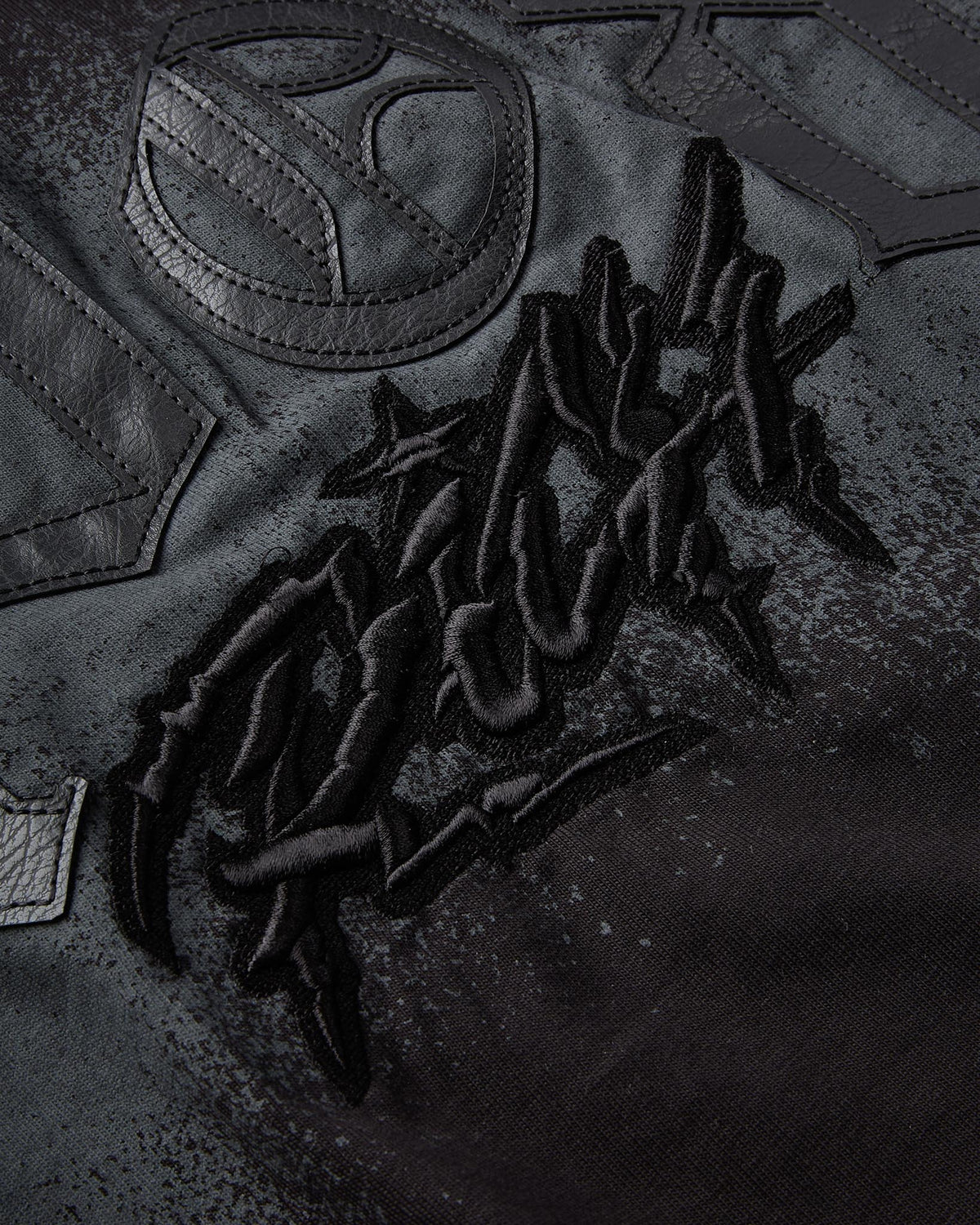 Close-up of a black leather jacket featuring a large, raised black embroidered graphic on the front and a circular leather patch.