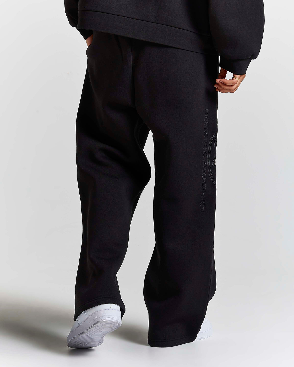 A person shown from the back wearing loose black sweatpants and a black sweatshirt, with white sneakers, against a light gray studio background.