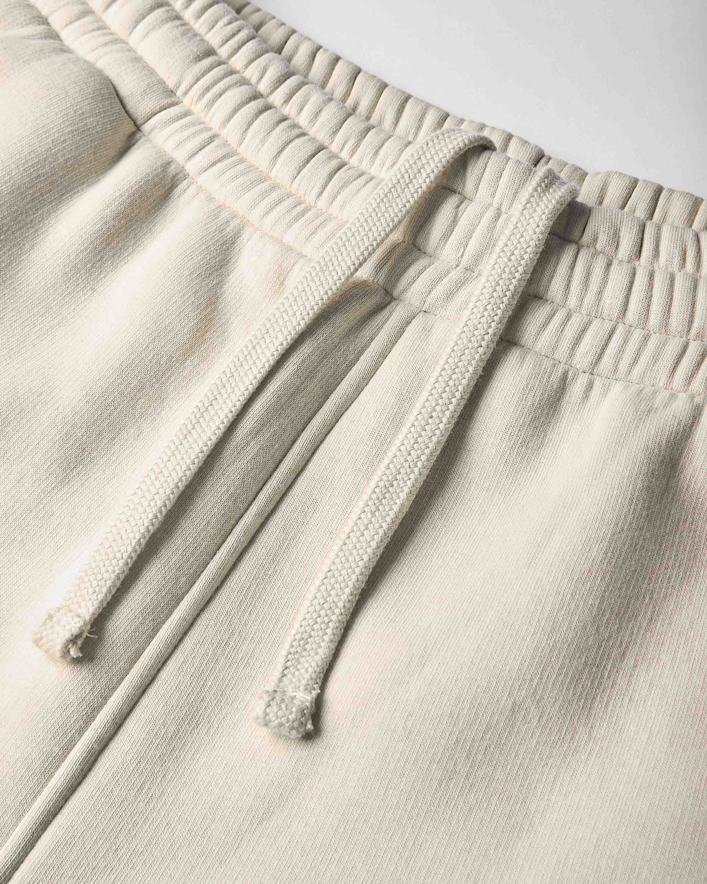 Close-up of a pair of light beige or cream sweatpants showing the textured fabric, elastic waistband, and matching drawstrings.