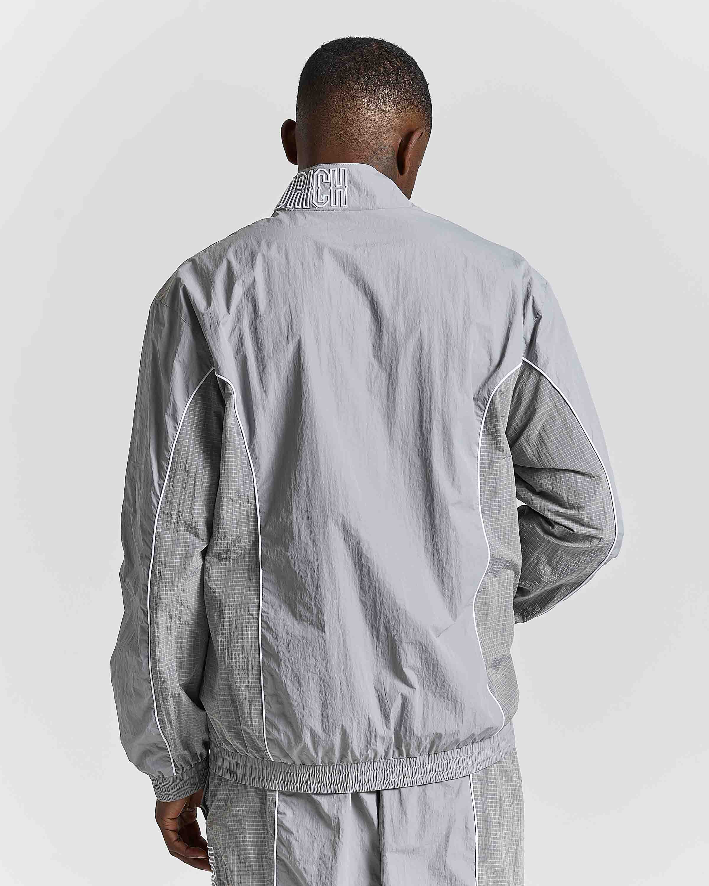 A man viewed from behind wearing a light gray windbreaker jacket with textured gray knit sleeves and a matching gray track pants, against a light gray studio background.