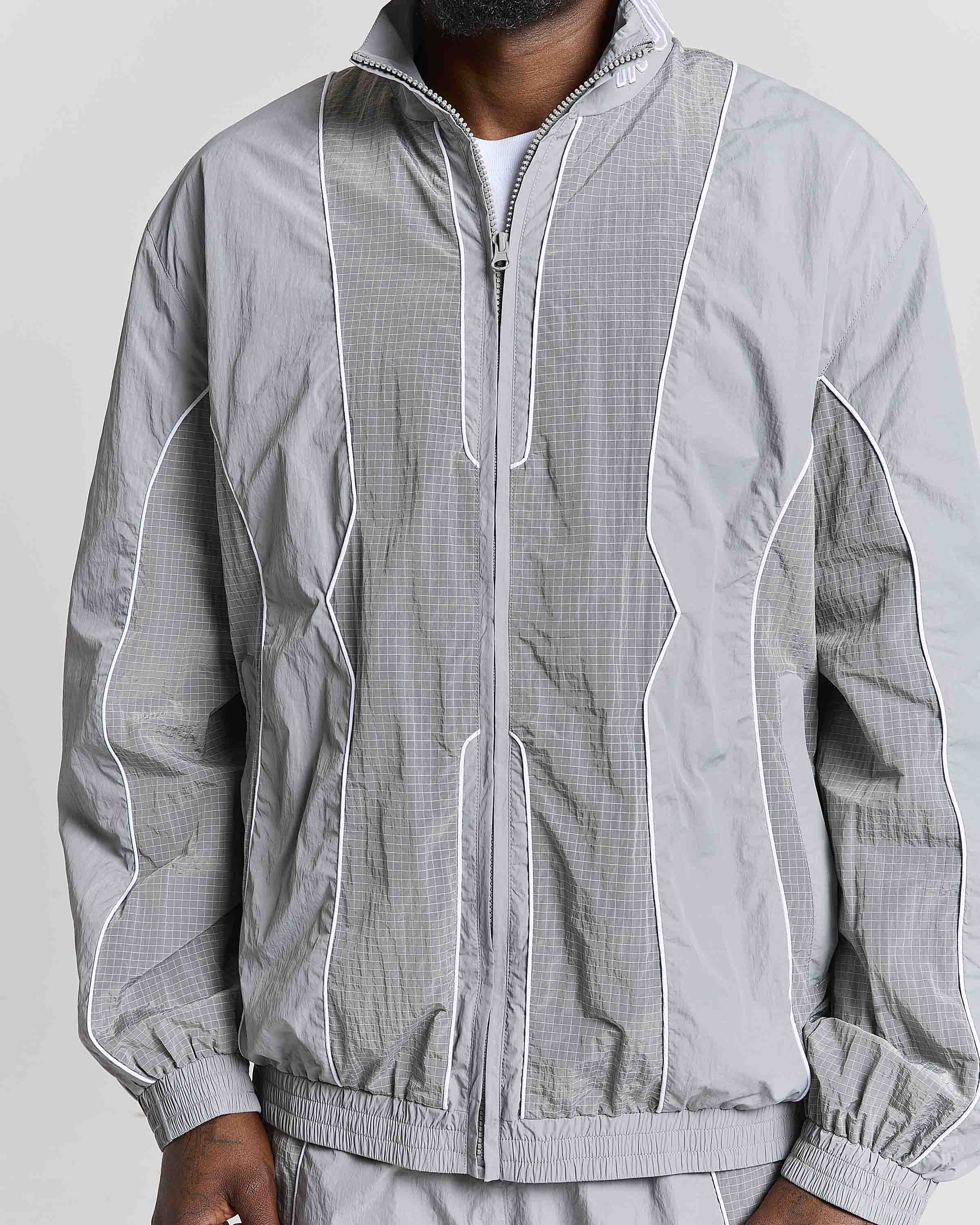 A close-up of a person wearing a light gray zip-up windbreaker jacket with white piping and a subtle grid texture, paired with matching gray pants against a plain light background.