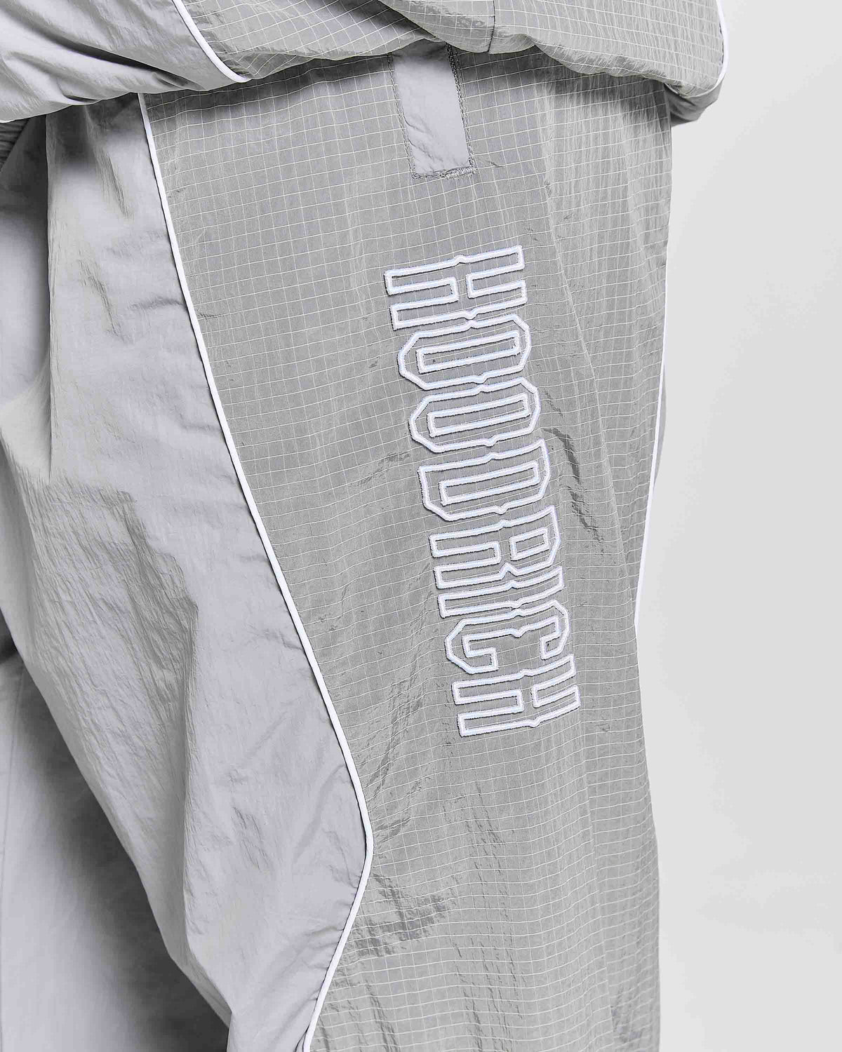 Close-up of light gray athletic pants with a white grid texture, white piping along the side seam, and a large white outlined vertical 'HOODRICH' logo on the leg.