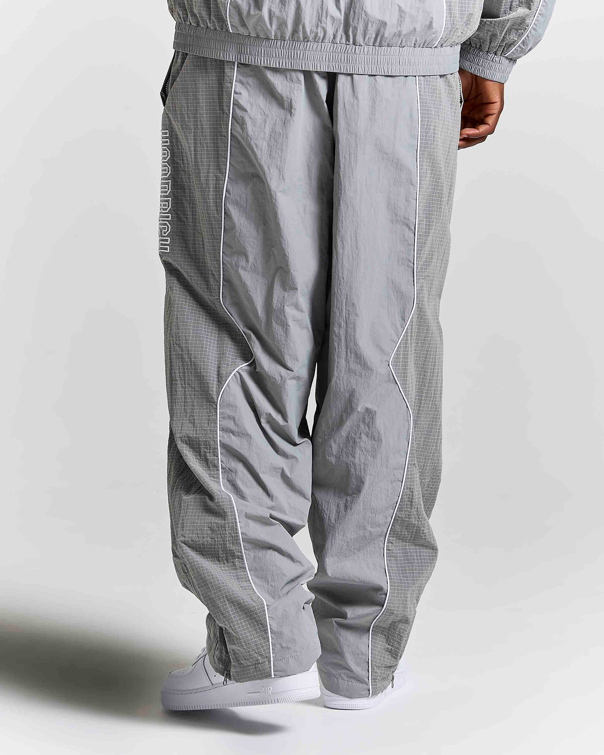 A person shown from the back wearing light gray wide-leg pants with white contrast piping and white sneakers, photographed against a plain light gray studio background.