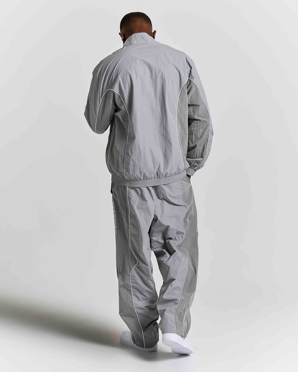 A man viewed from behind wearing a light gray windbreaker jacket and matching light gray pants, standing against a plain light gray studio background.