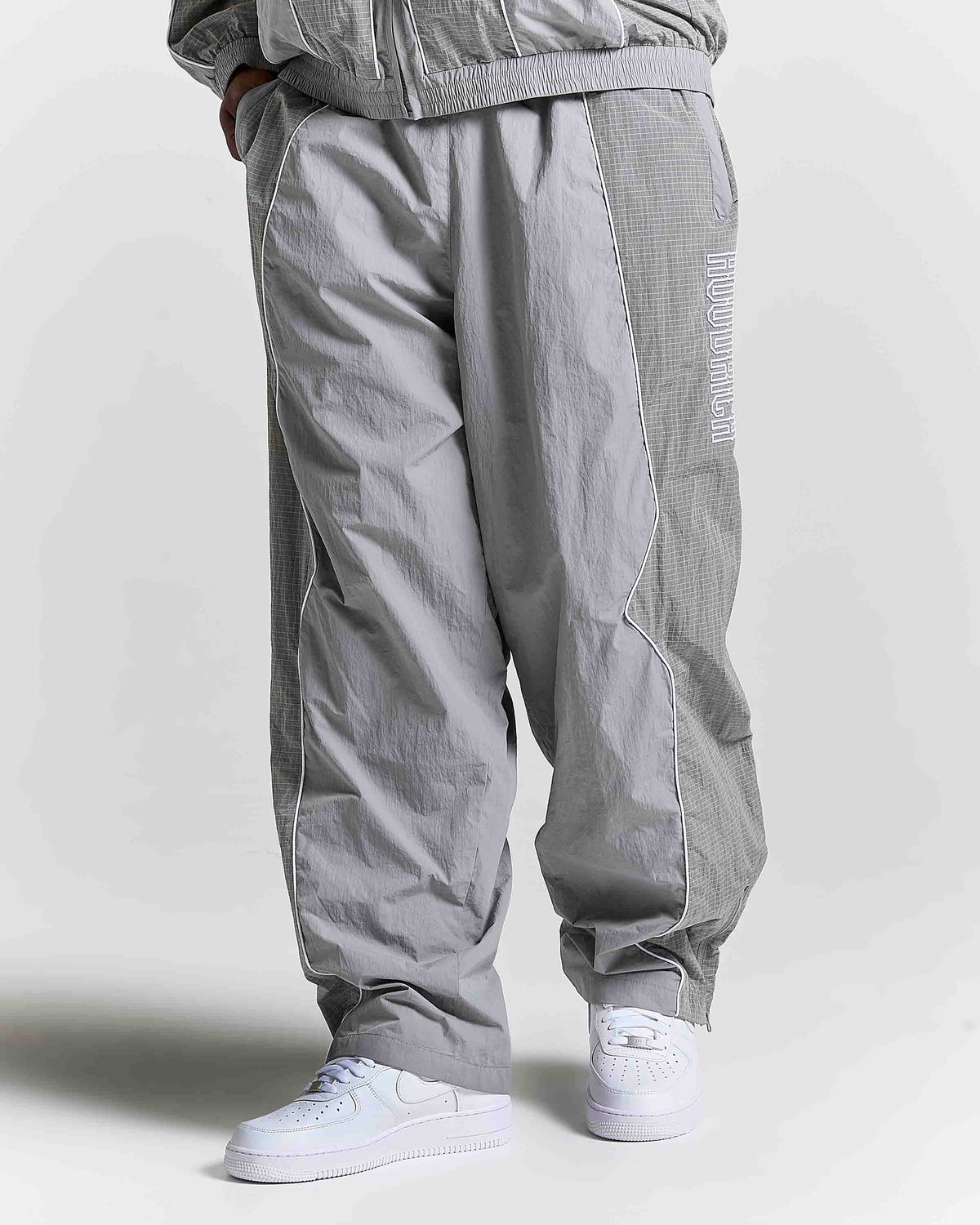 A person wearing loose-fit light gray jogger-style pants with a subtle grid texture, side pockets, and white sneakers against a plain light gray studio background.