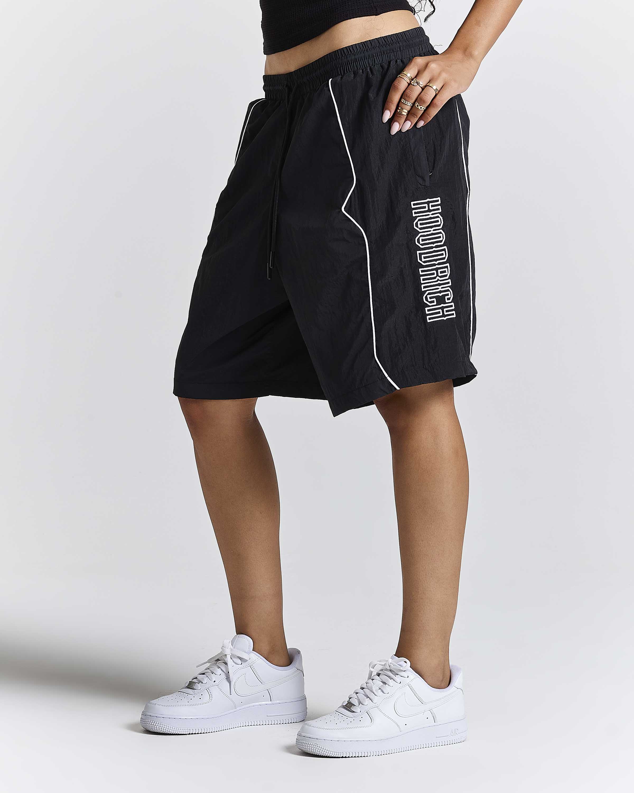 A person wearing black athletic shorts with white piping and vertical white 'HOODRICH' text on the side, paired with white sneakers, photographed against a light gray studio background.