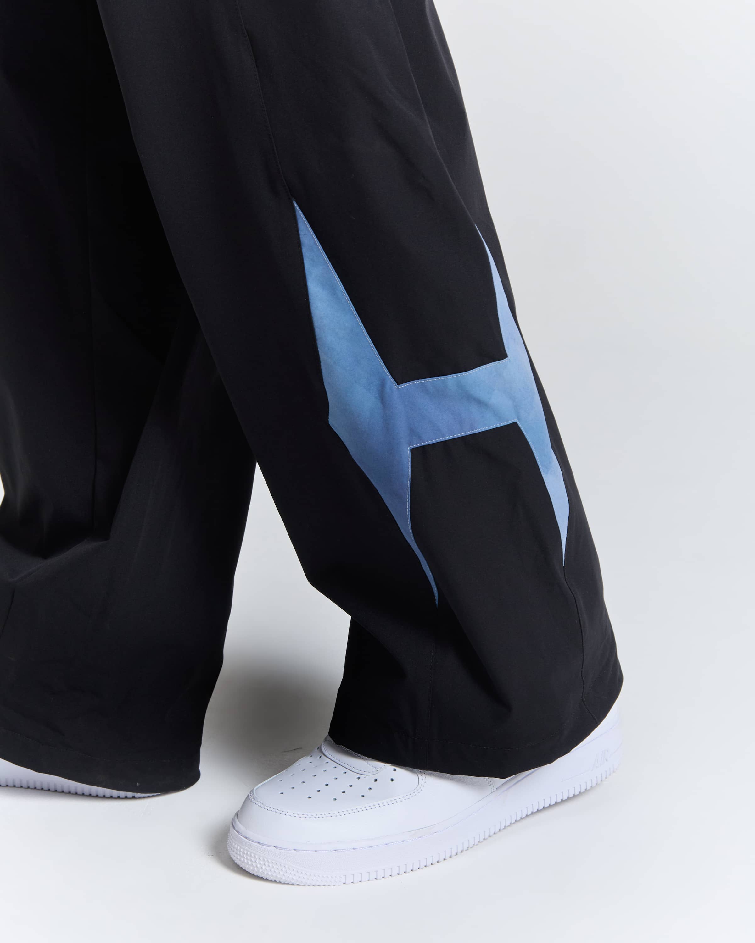 Close-up of a person's lower legs wearing black wide-leg pants with a large light blue geometric star graphic on the outer side and white low-top sneakers, against a light gray studio background.