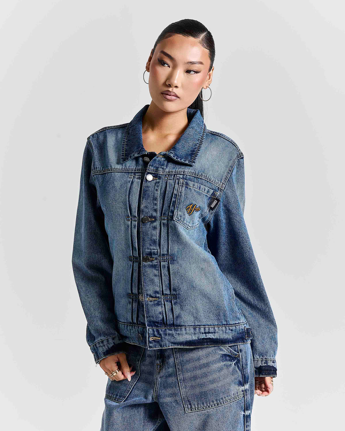 A model wearing a blue denim jacket with a collar, button front, and a small embroidered logo on the chest pocket, paired with matching blue denim pants against a light gray studio background.
