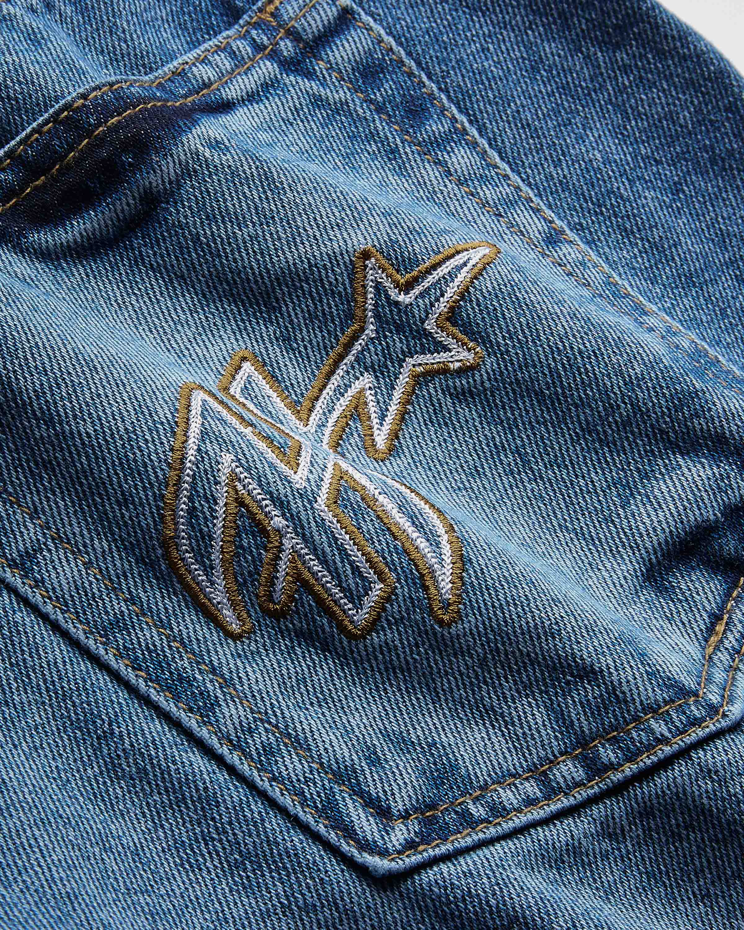 Close-up of a blue denim jacket showing a pocket with an embroidered white and gold monogram logo.