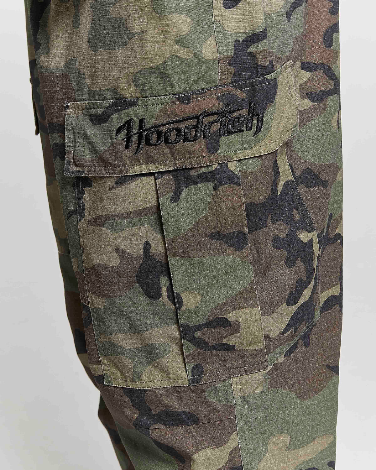 Close-up of camouflage cargo pants featuring a large flap pocket with 'Hoodrich' embroidered in black script on the tab, set against a plain light gray background.