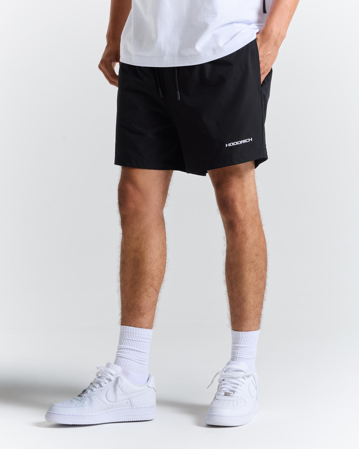 A person wearing black athletic shorts with a small white 'HOODRICH' logo on the lower left leg, paired with a white t-shirt, white socks, and white sneakers, shown from the waist down on a light gray studio background.