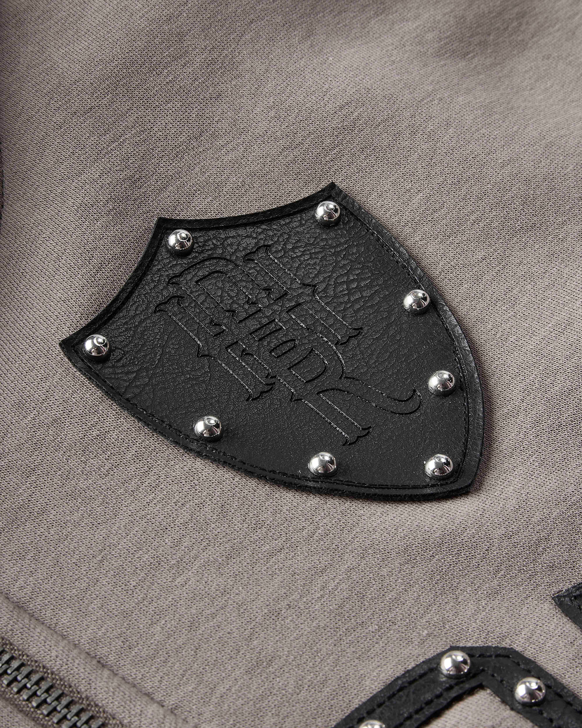 Close-up of a black shield-shaped leather patch with silver studs and an embossed logo, attached to a taupe-gray textured fabric garment.