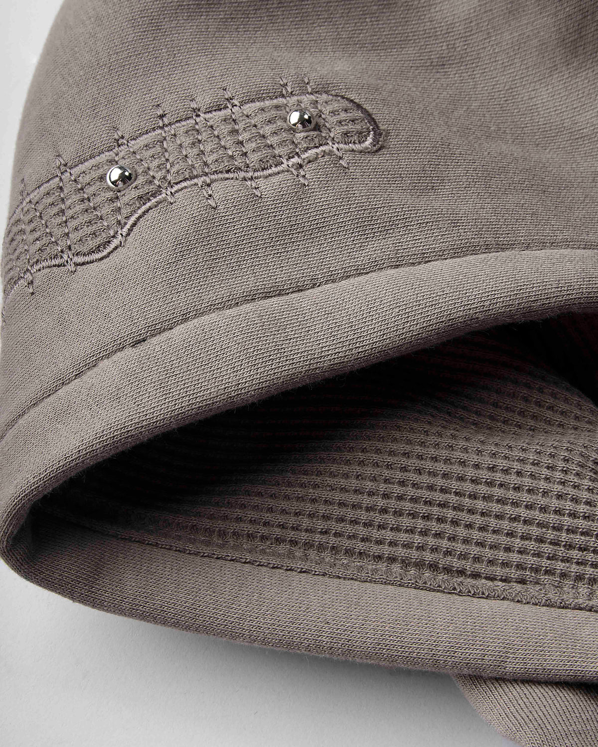 Close-up of a taupe gray fabric accessory, showing a waffle-knit texture, stitched detail with two small silver rhinestones, and a folded edge.
