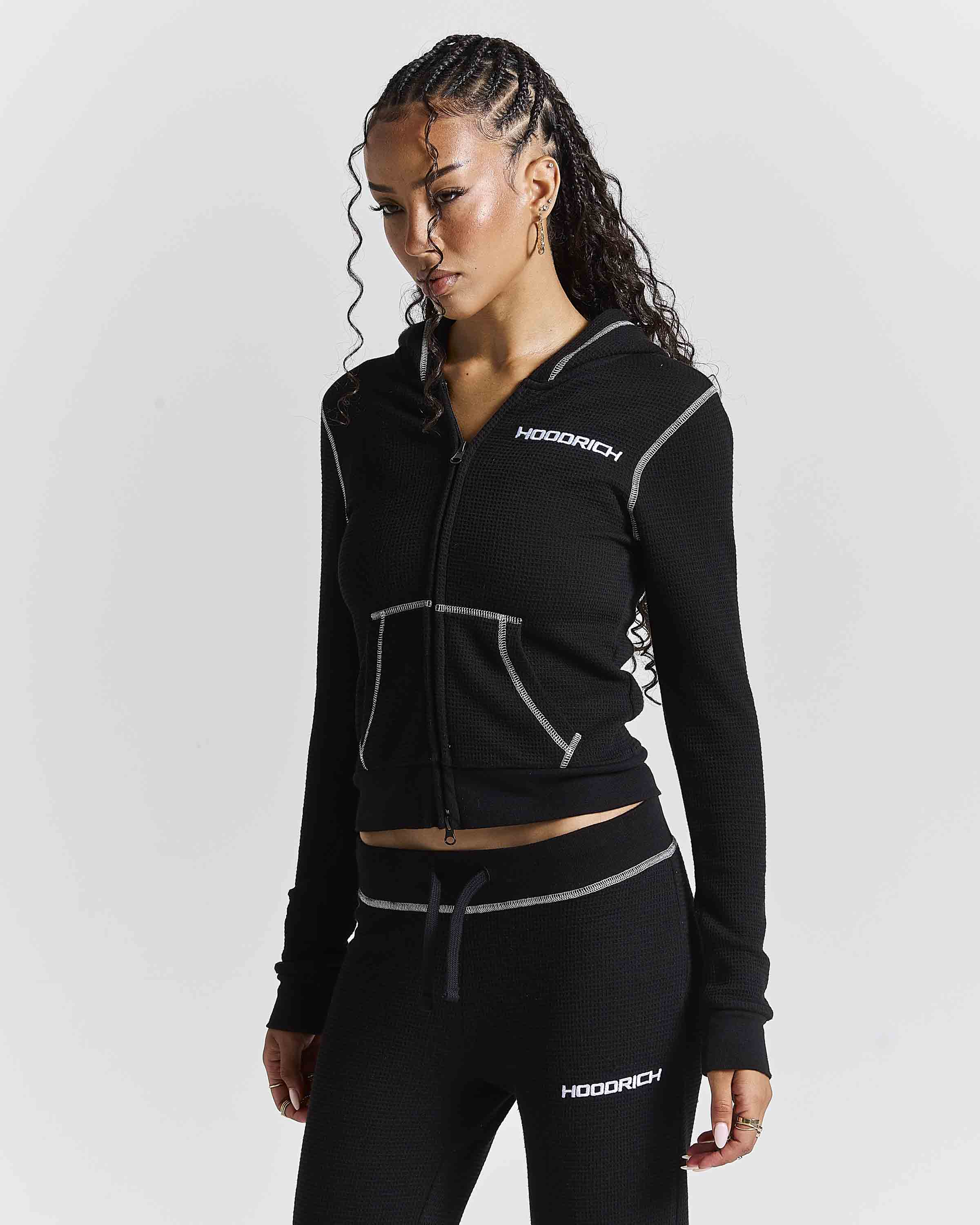 A woman modeling a black zip-up hoodie and matching black joggers with white contrast stitching and white HOODRICH logos, photographed against a light gray studio background.