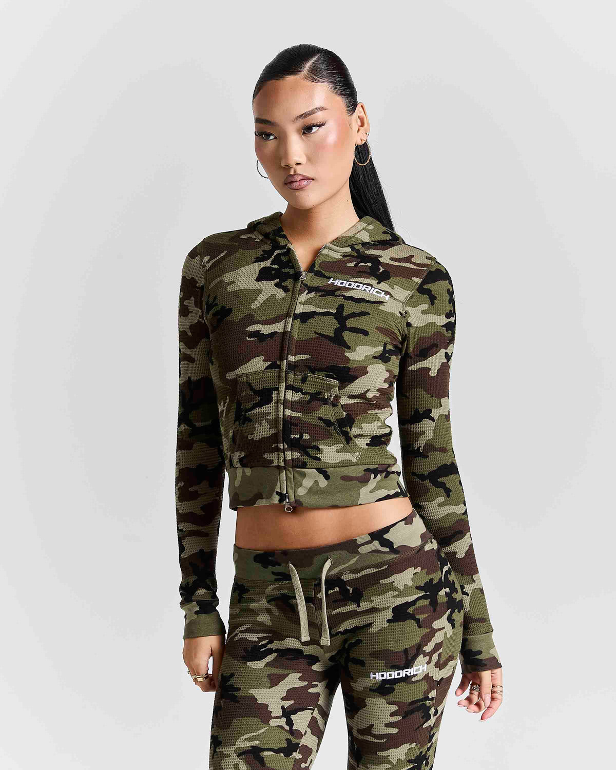 A model wearing a matching camouflage zip-up hoodie and jogger pants set with white HOODRICH branding, photographed against a light gray studio background.