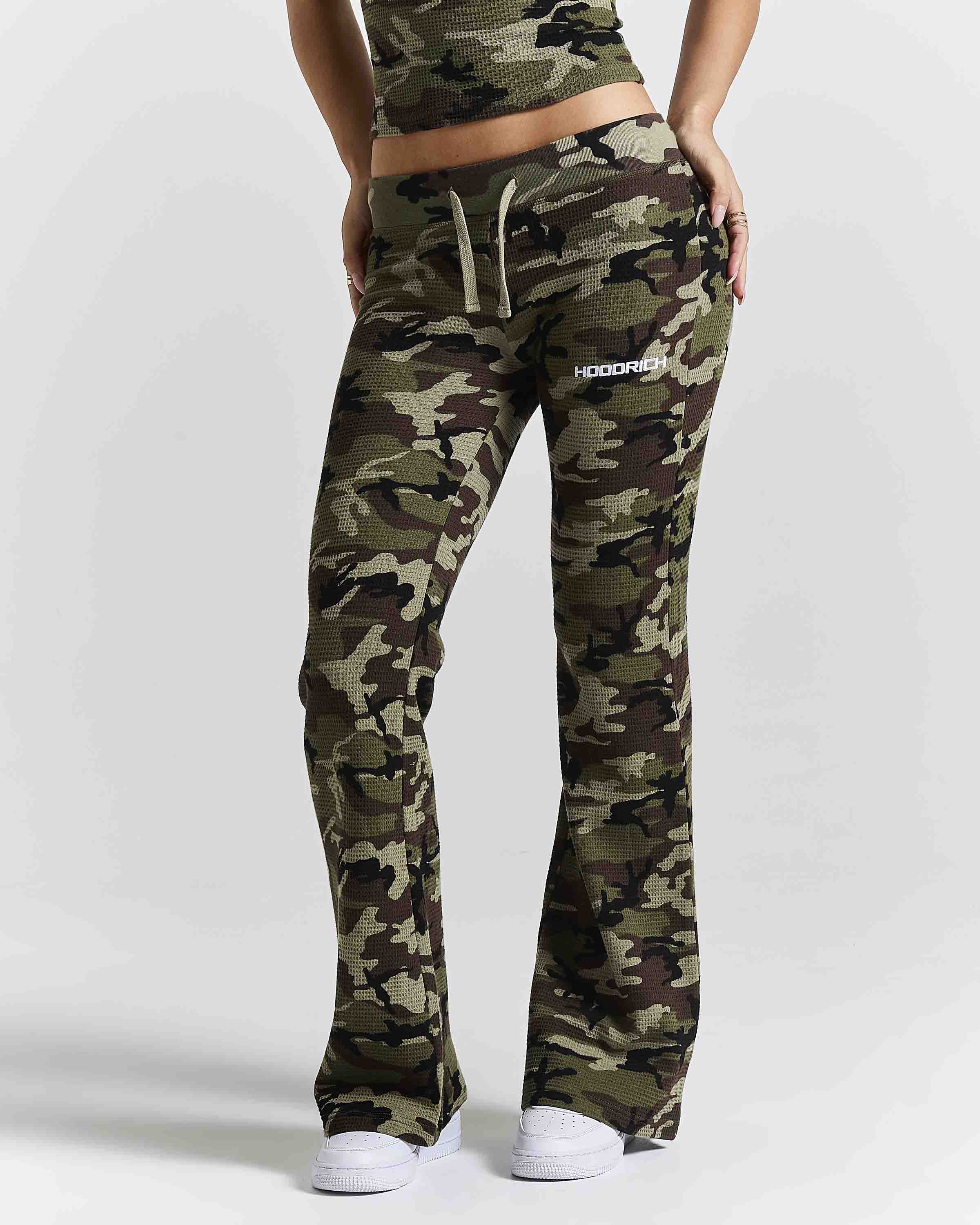 A model wearing camouflage sweatpants with a drawstring waist and a white 'HOODRICH' logo on the left hip, paired with a matching camouflage top and white sneakers, against a light gray studio background.