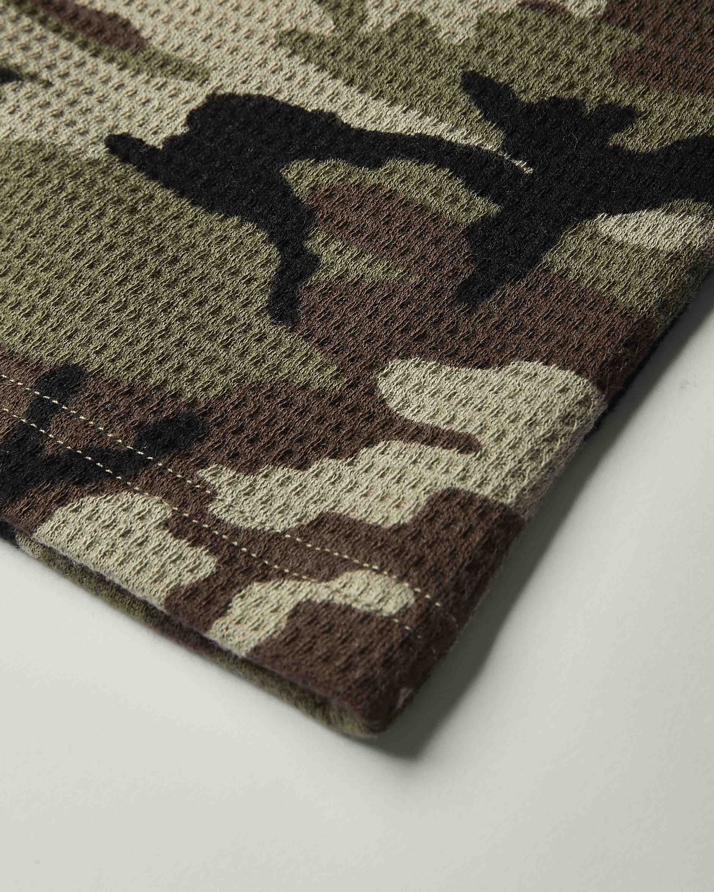 Close-up of a folded waffle-knit fabric with a classic camouflage pattern in olive green, brown, black, and beige, showing the textured knit construction and neat stitching.