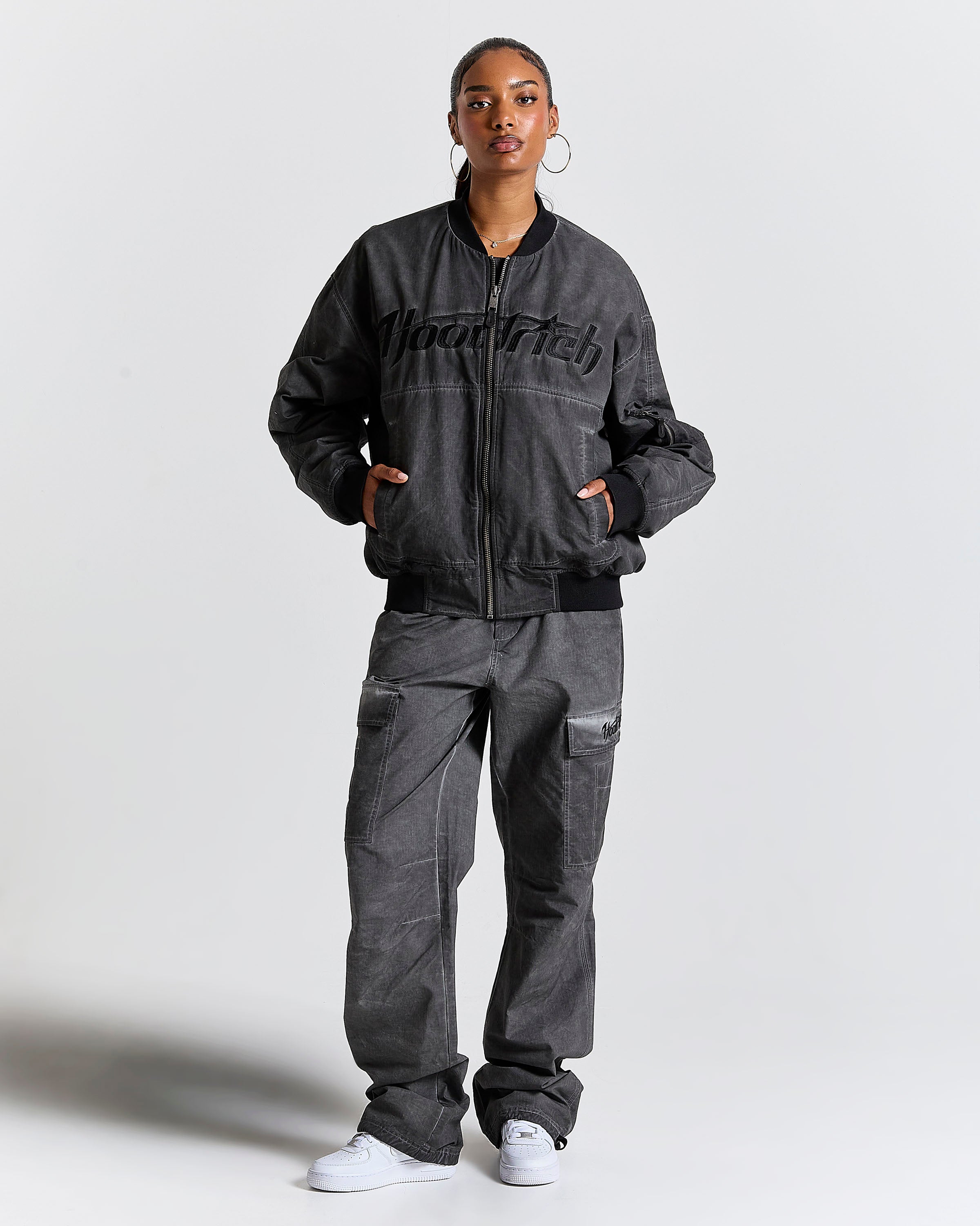 Person wearing a dark gray bomber jacket and pants with 'Hoodrich' branding on a light gray background