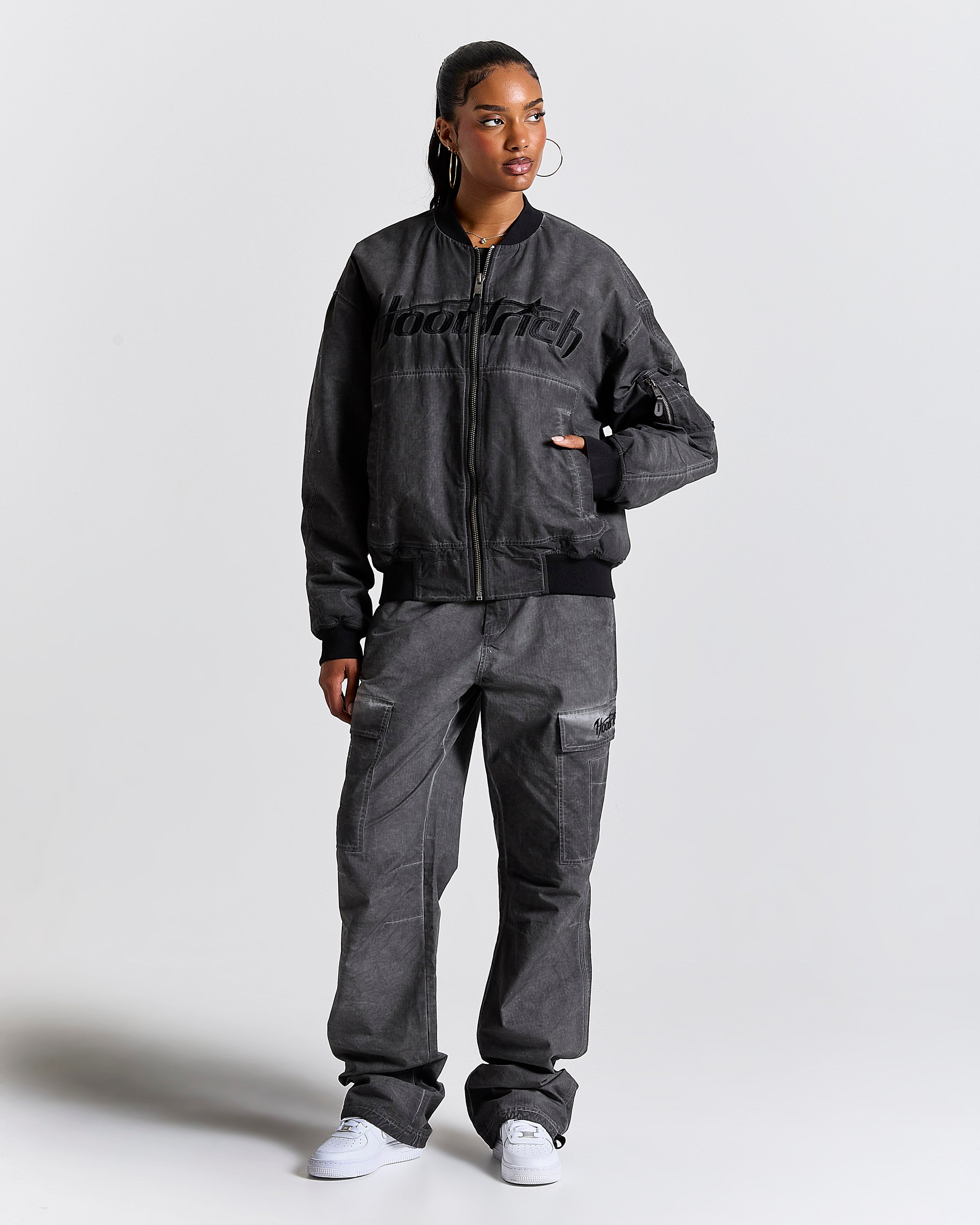 A model wearing a dark gray washed bomber jacket with 'HOODRICH' printed across the chest, matching dark gray cargo pants, and white sneakers, standing against a light gray studio background.