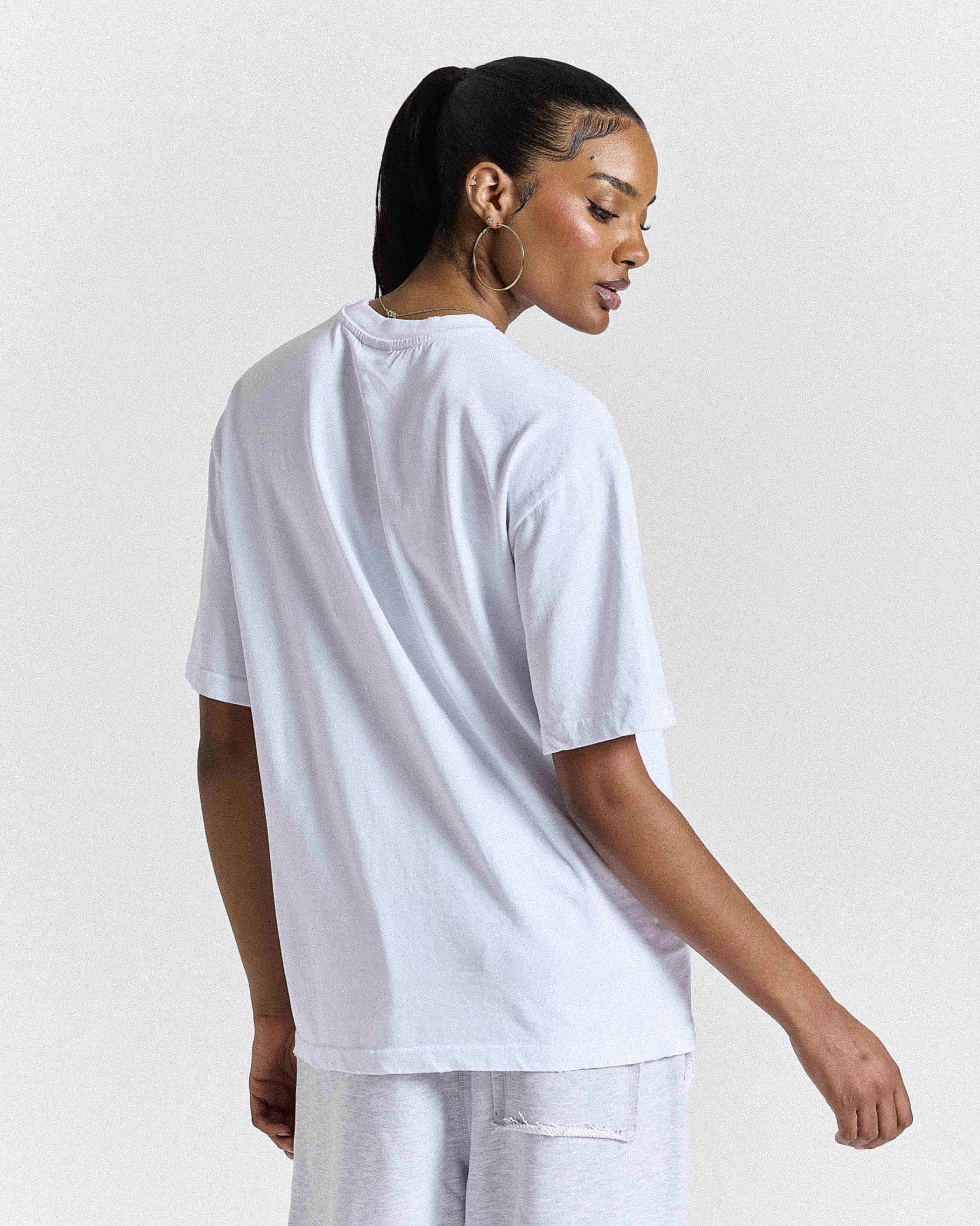 A woman shown from the back wearing an oversized white short-sleeve t-shirt and light gray sweatpants against a plain light gray studio background.