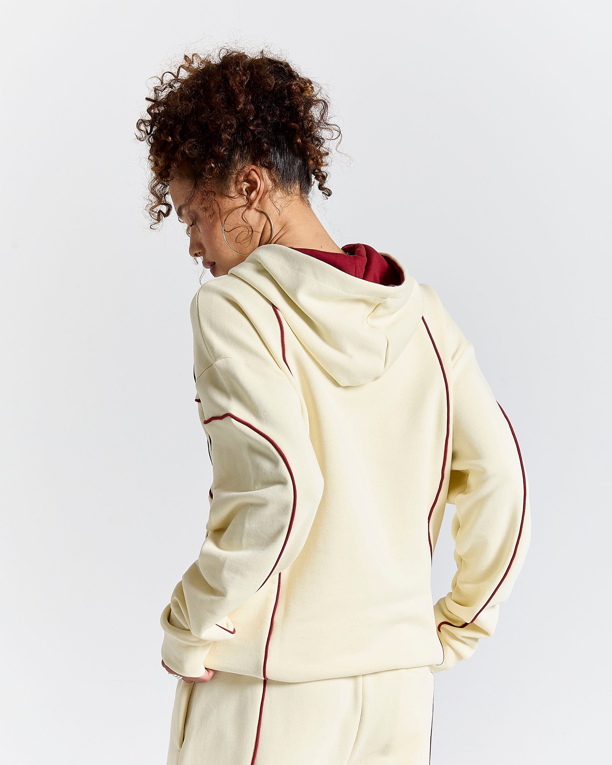 A model shown from the back wearing a cream-colored hoodie with burgundy piping and lining, paired with matching sweatpants, against a light gray studio background.