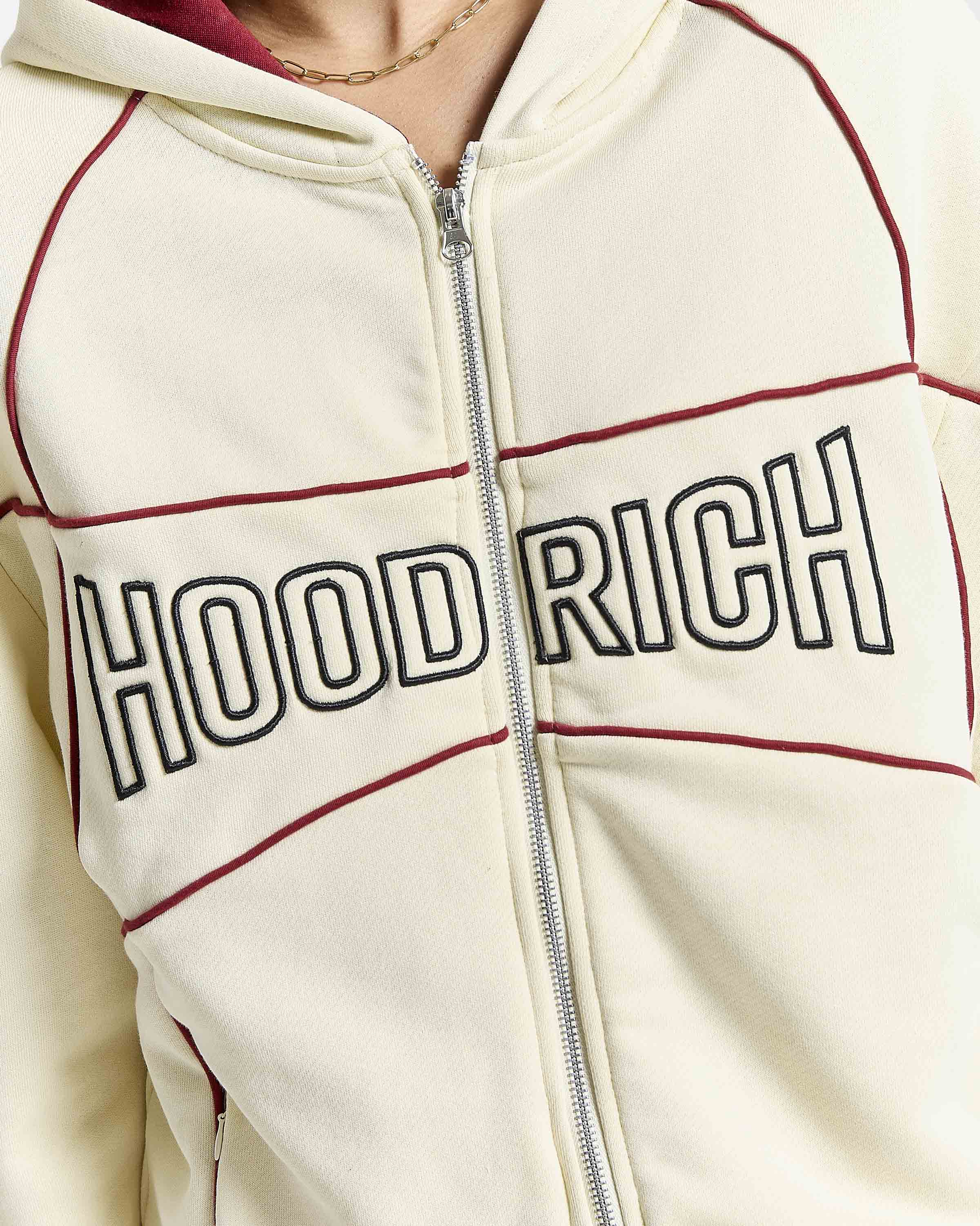 Close-up of a cream zip-up hoodie with 'HOODRICH' embroidered across the chest in black outlined letters, accented by dark red piping lines.