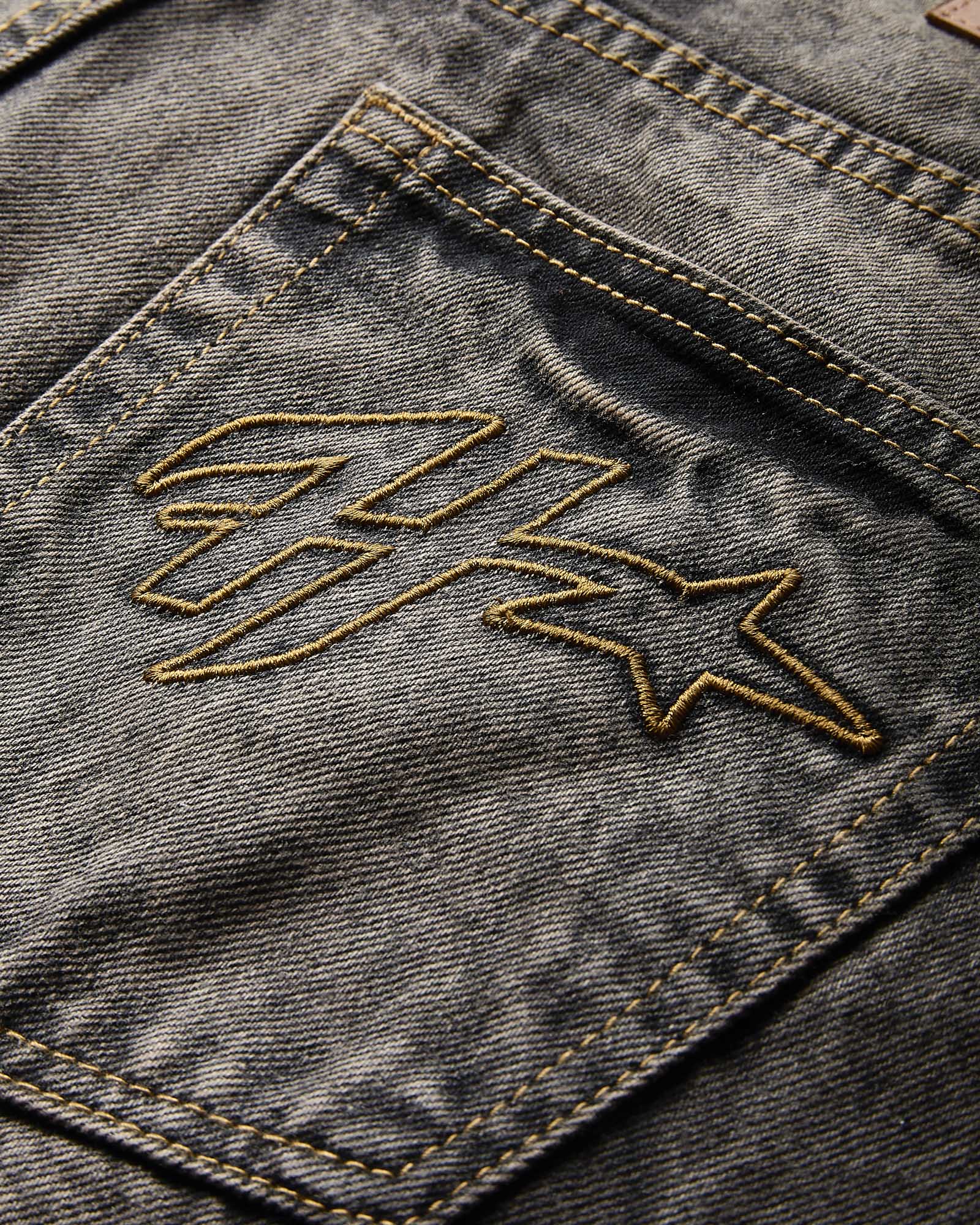Close-up of a dark gray washed denim pocket with gold contrast stitching and a gold embroidered logo.