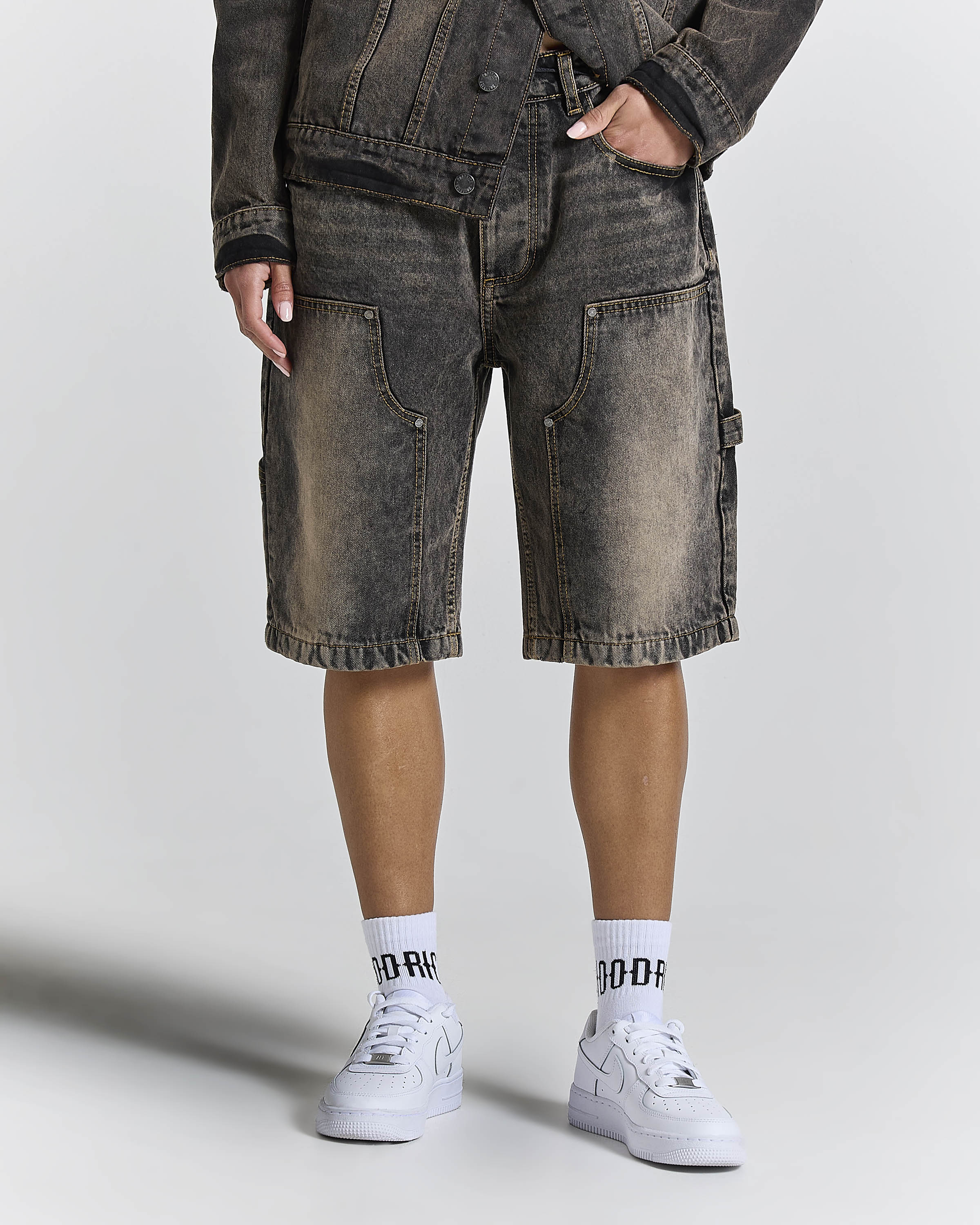 A person wearing washed charcoal gray denim cargo shorts, a matching dark jacket, white socks with black lettering, and white sneakers, photographed against a light gray studio background.