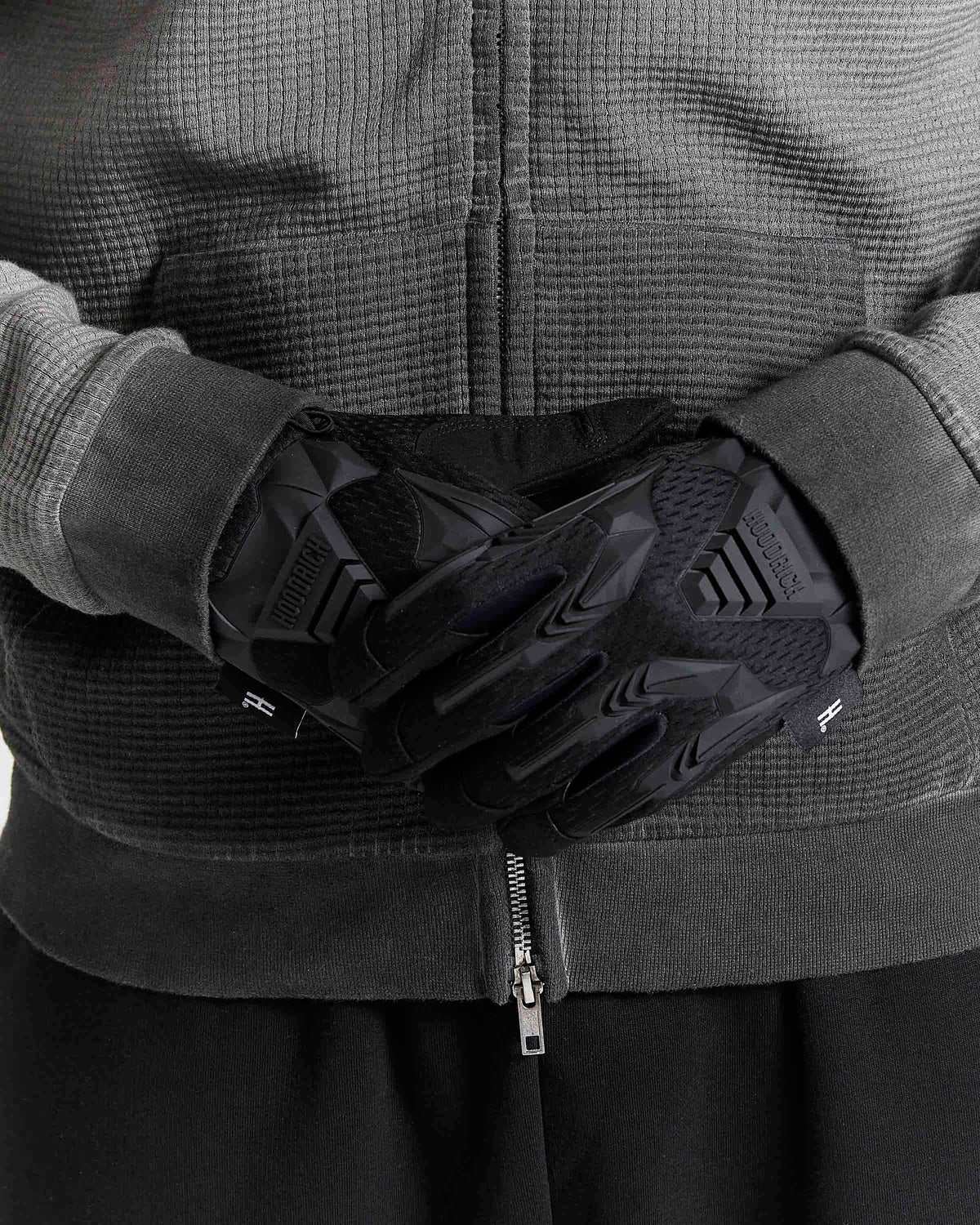 Black protective gloves worn on a person's hands, shown against a dark gray textured zip-up jacket.