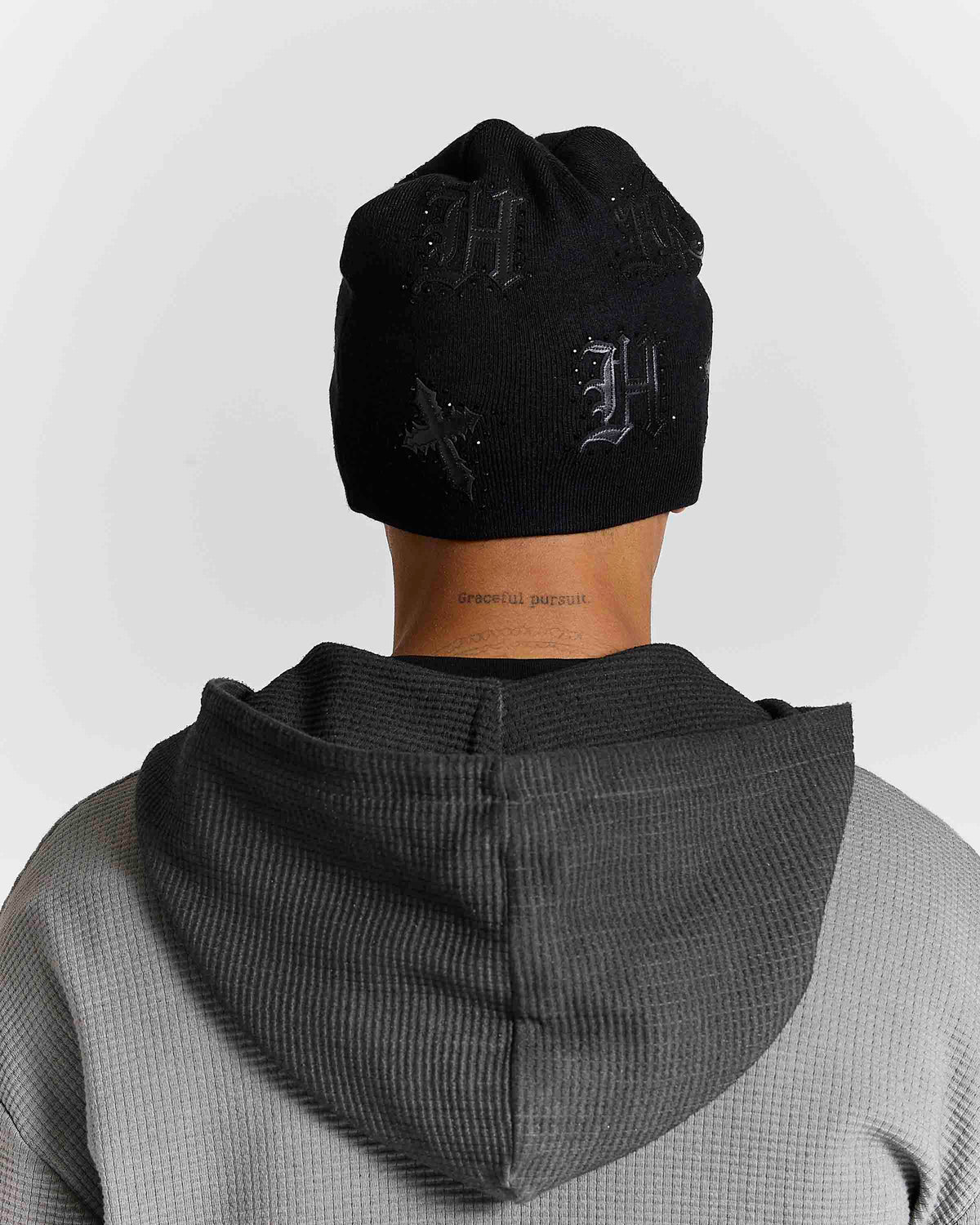 A person shown from the back wearing a black knit beanie with tonal embroidered gothic lettering and a dark gray ribbed hooded sweatshirt against a light gray studio background.