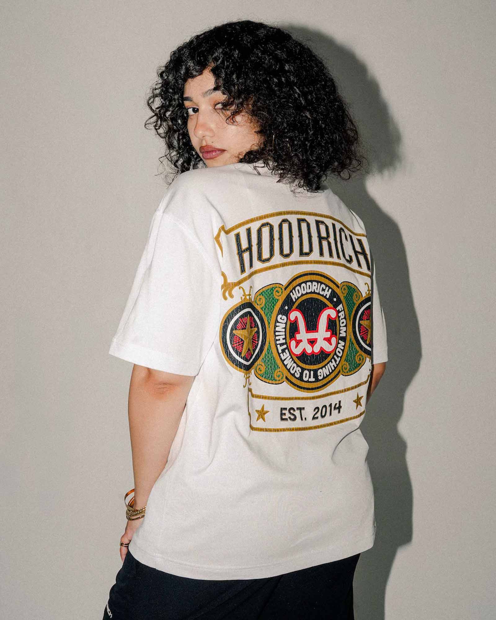 A model with curly black hair is shown from the back wearing a white HOODRICH t-shirt with a large graphic print on the back, paired with black pants, set against a plain gray wall.