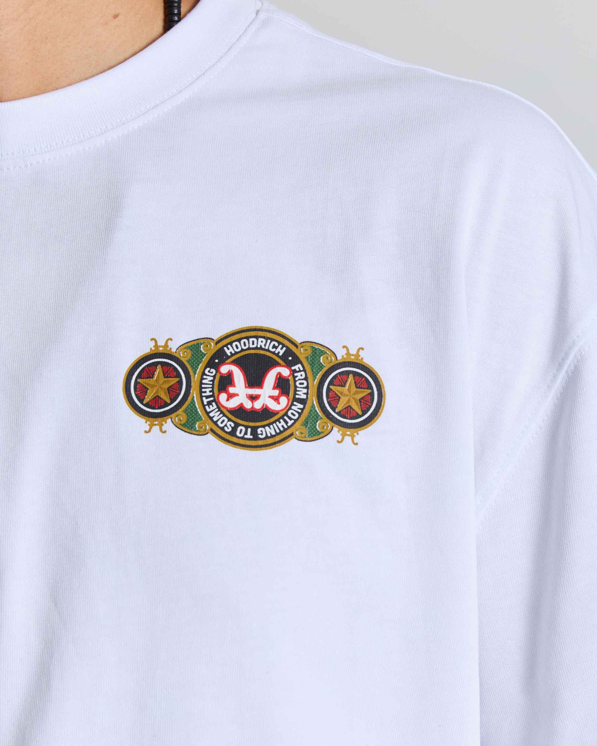 A close-up of a white long-sleeve t-shirt featuring a colorful embroidered chest graphic. The graphic displays a central black oval with a red and white stylized '365' logo, flanked by two gold star emblems and the words 'HOODRICH' and 'ESTD 2014'.