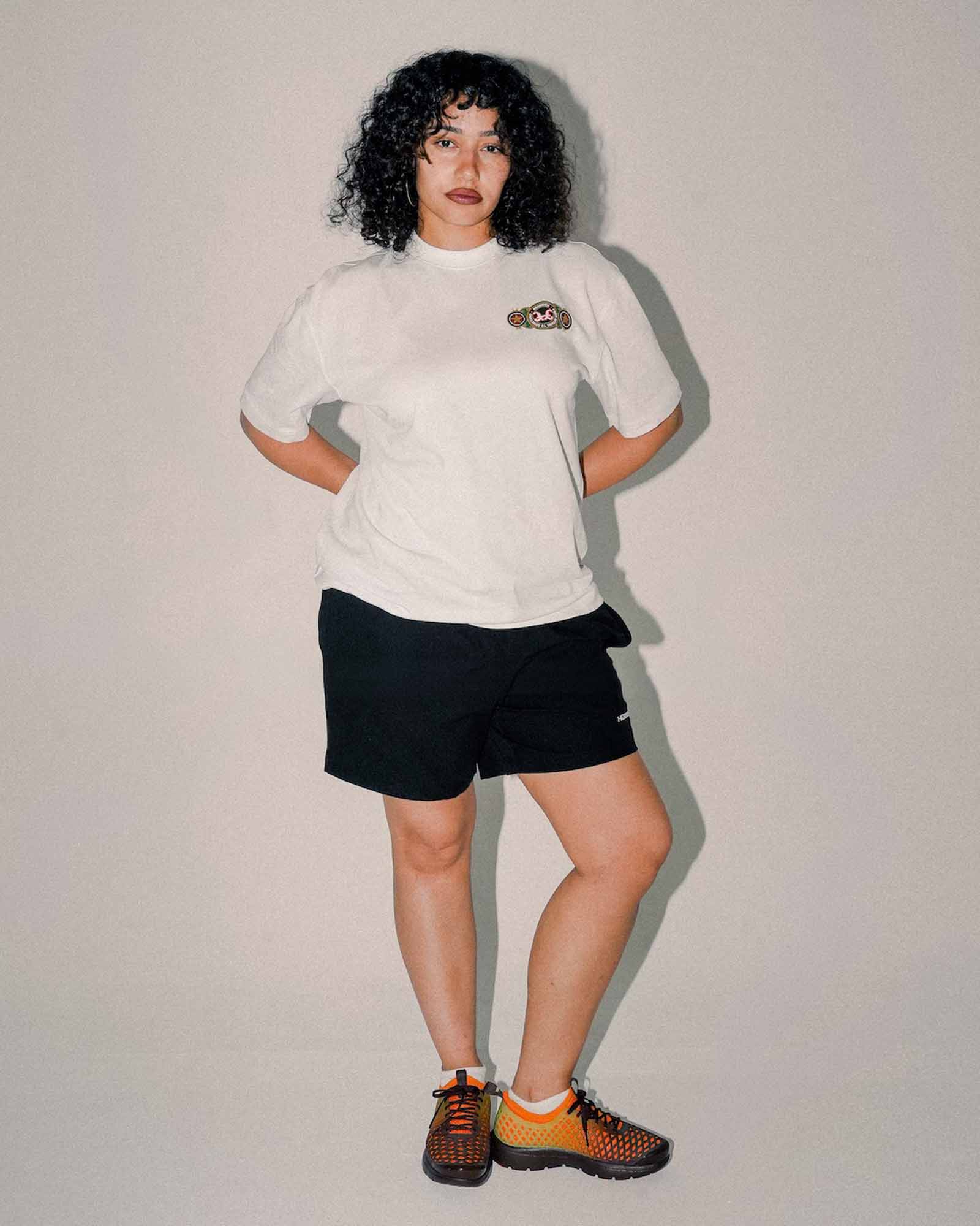 A model with curly black hair stands against a light gray studio background wearing a white t-shirt with a small graphic on the left chest, black athletic shorts, and orange and black sneakers.