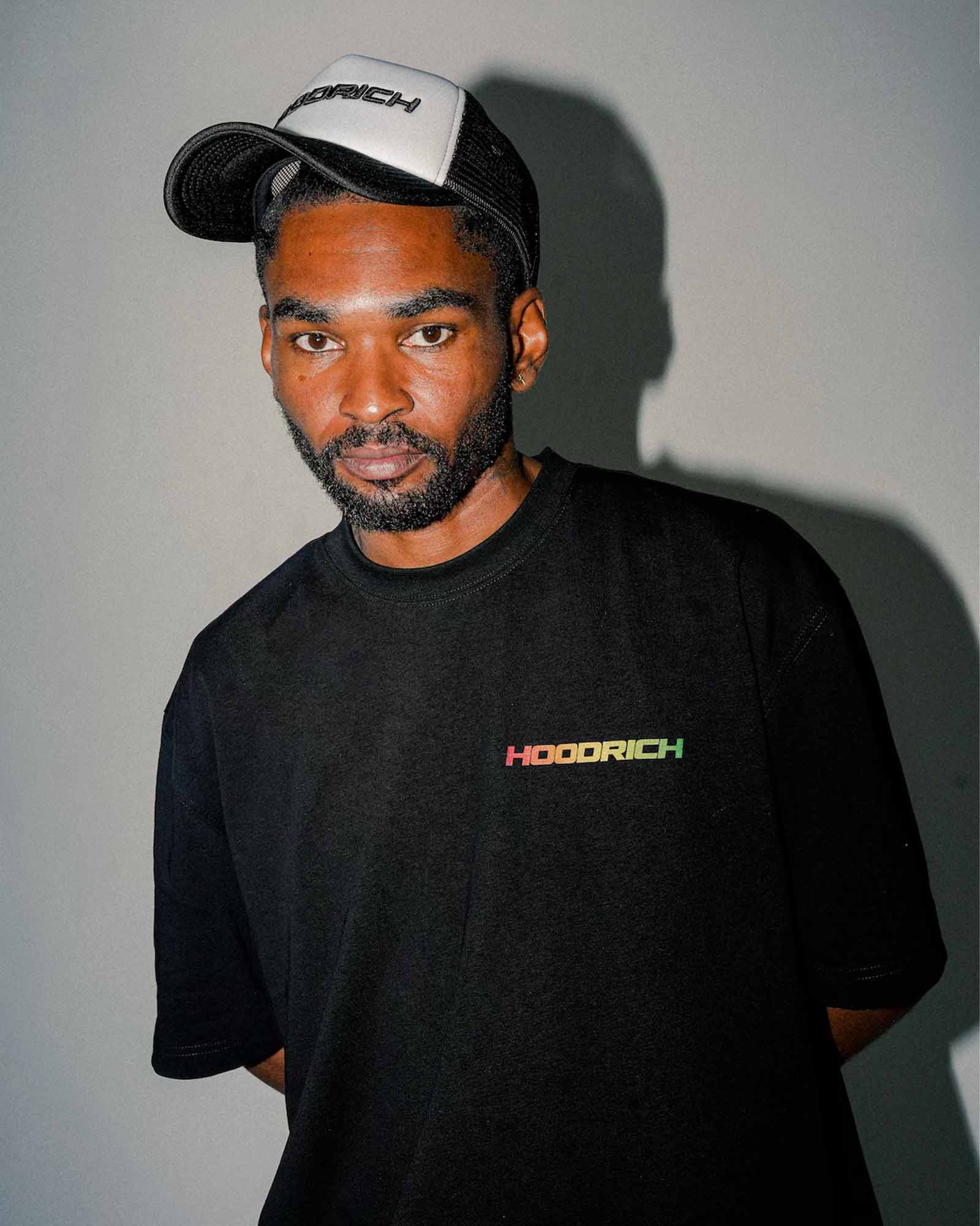 A man wearing a black HOODRICH t-shirt and a black and white trucker hat, photographed against a plain gray wall with a strong shadow.