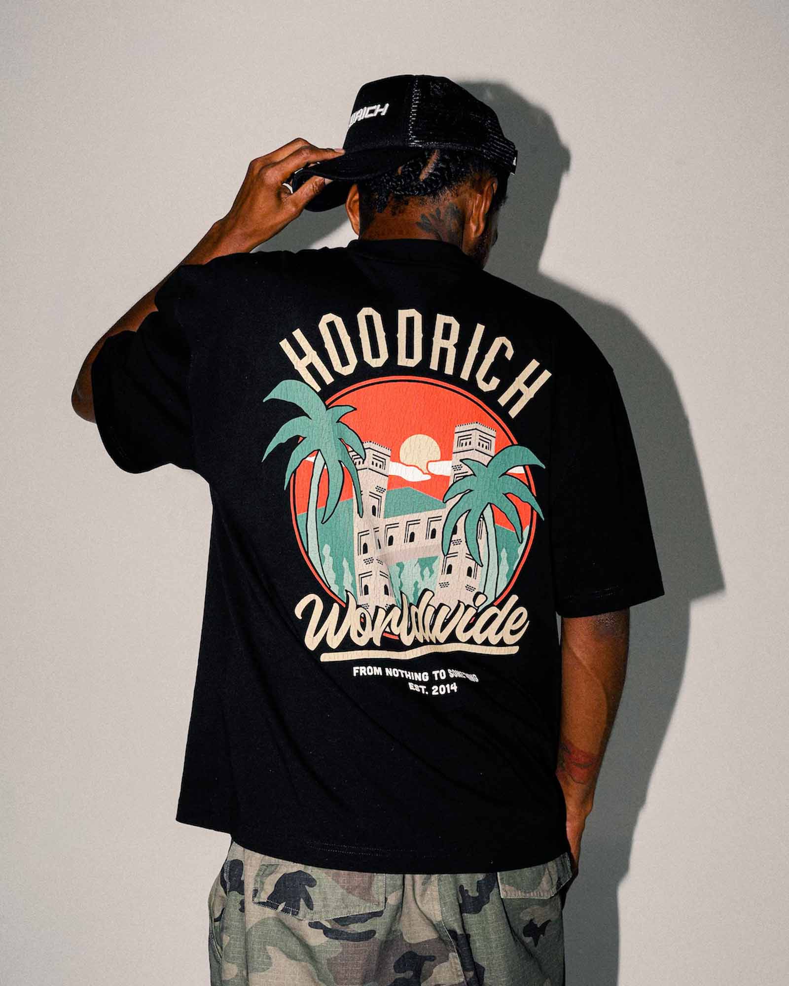 A man viewed from behind wearing a black HOODRICH Worldwide t-shirt with a large retro graphic on the back, paired with camouflage shorts and a black cap, against a plain light gray wall.