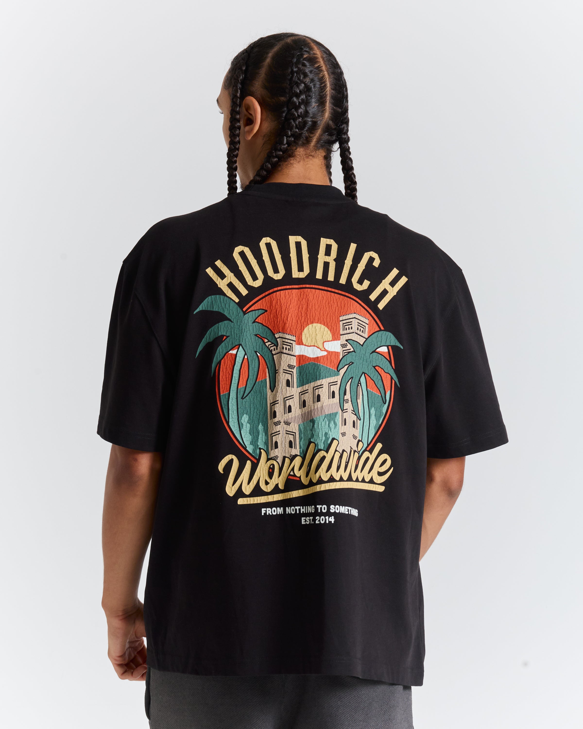 A person shown from the back wearing a black short-sleeve graphic T-shirt. The shirt features a large retro-style print with the words 'HOODRICH' and 'Worldwide' in tan lettering, along with a circular sunset scene, palm trees, and a building.