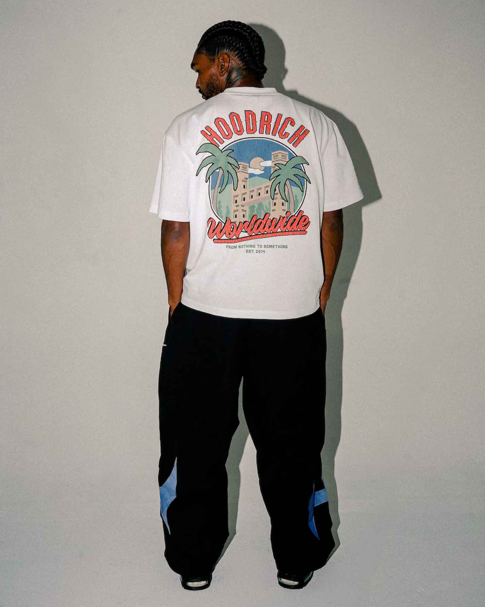 A man stands with his back to the camera wearing a white t-shirt with a large graphic print on the back that reads 'HOODRICH Worldwide' and black sweatpants with blue accents.