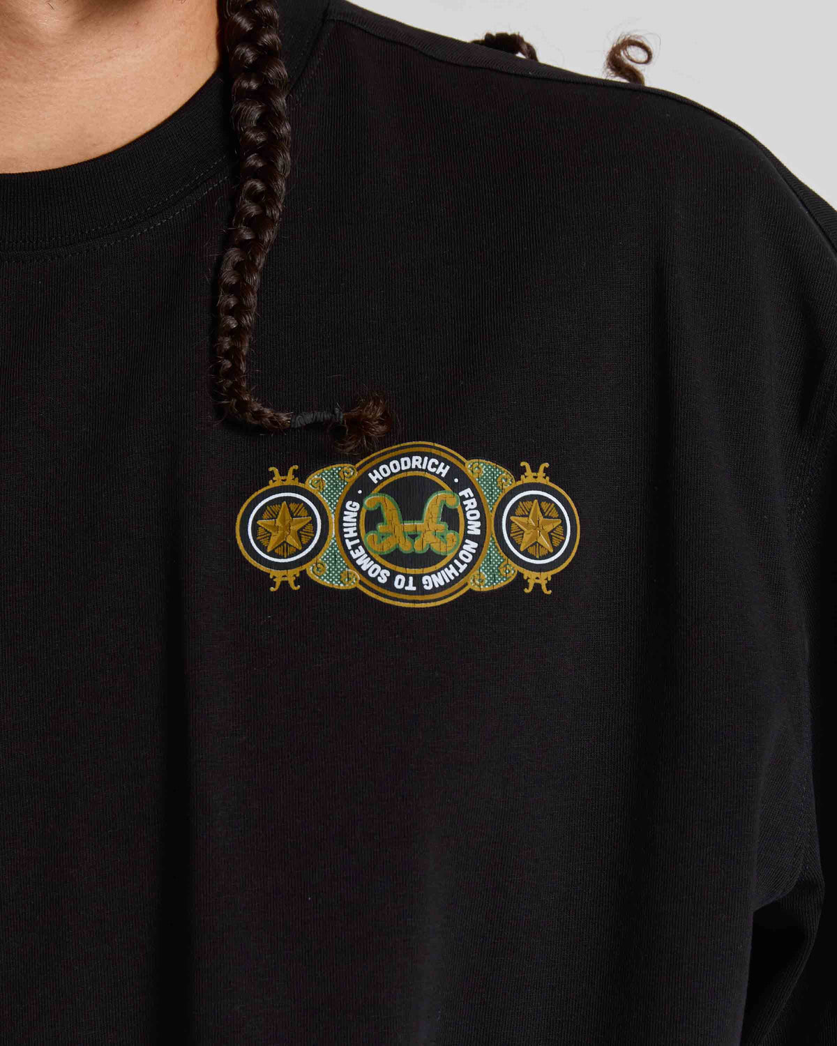 Close-up of a person wearing a black oversized T-shirt with a decorative chest graphic featuring gold, green, and white text and emblem details. A braided hair strand falls over the shirt.