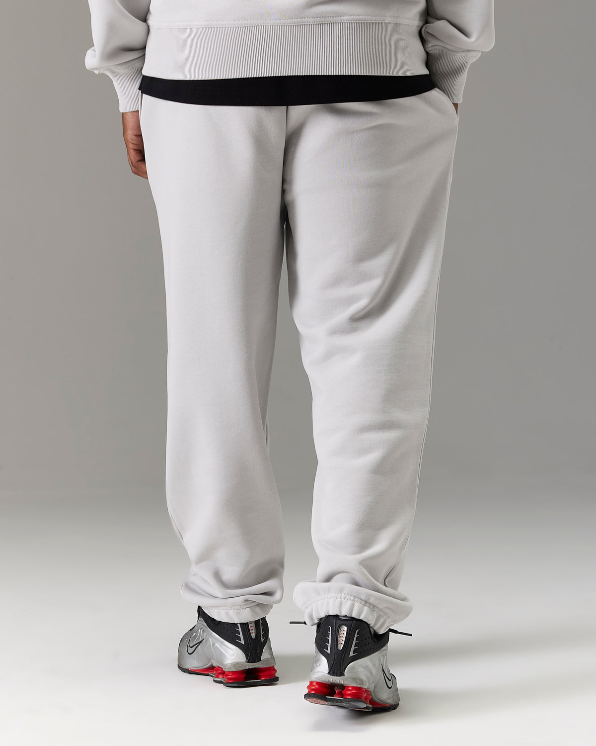 A person shown from the back wearing light gray sweatpants with a black waistband, a light gray sweatshirt, and gray sneakers with red accents, against a neutral gray studio background.