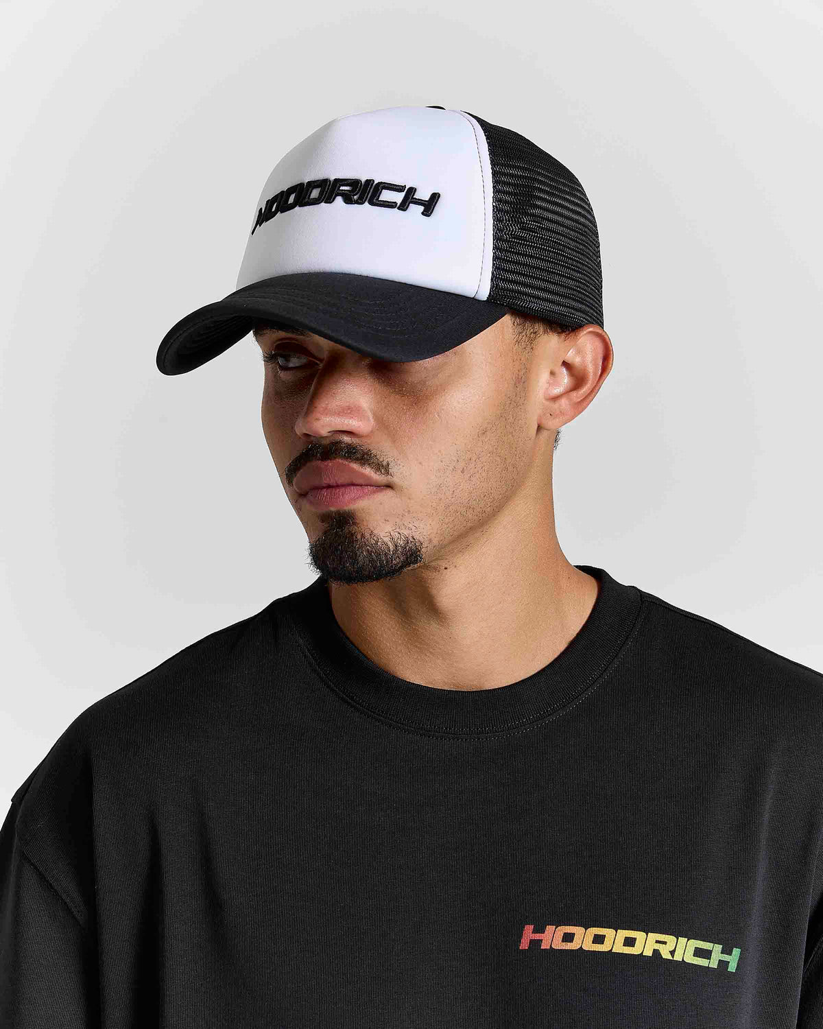 A man wearing a black and white trucker hat with 'HOODRICH' printed on the front, paired with a black hoodie featuring a small multicolor 'HOODRICH' logo on the chest, set against a light gray background.
