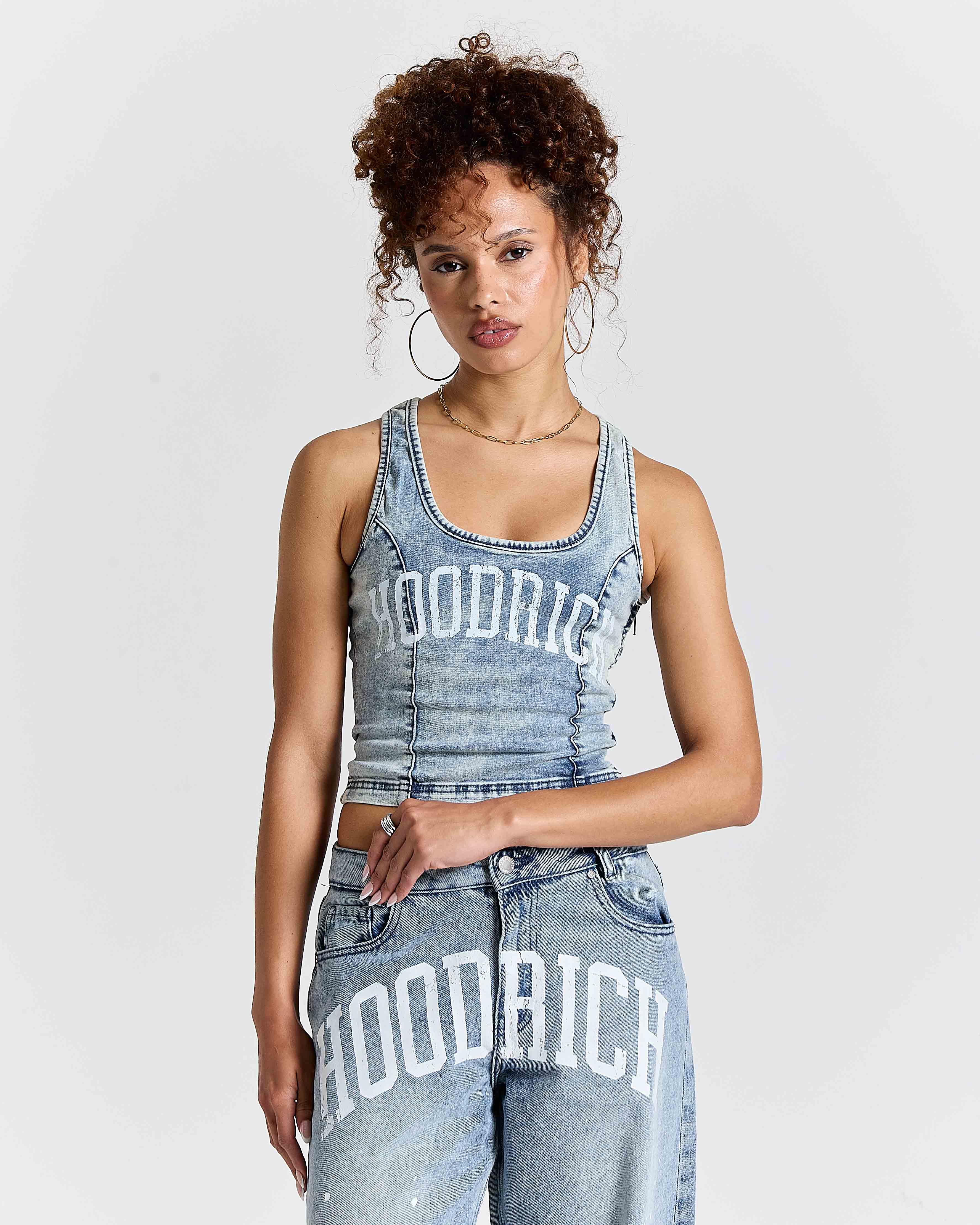 Woman wearing a denim tank top and jeans with 'HOODRICH' branding on a plain background