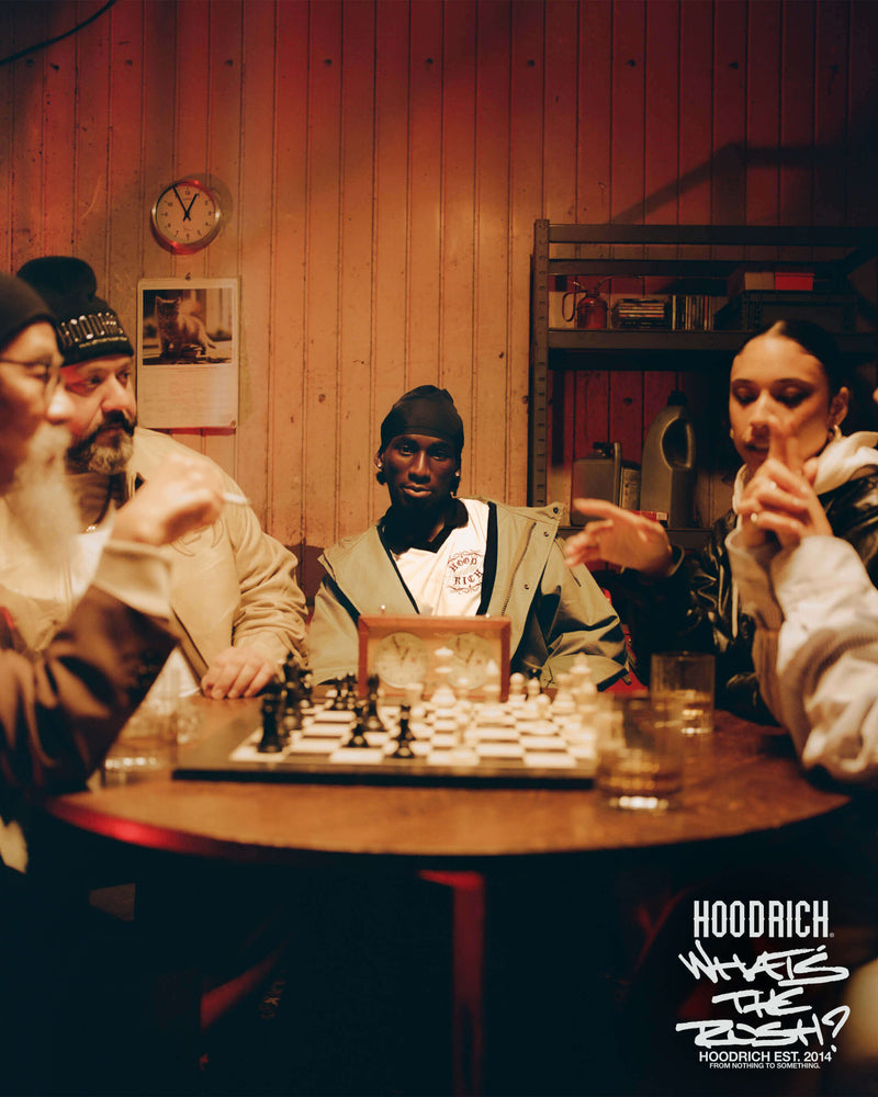 A group of people gathered around a wooden table in a dimly lit room with wood-paneled walls, with a chess game in progress. The image has a warm, moody orange-brown lighting and features the text 'HOODRICH' and 'WHAT'S THE RUSH' in the bottom right corner.