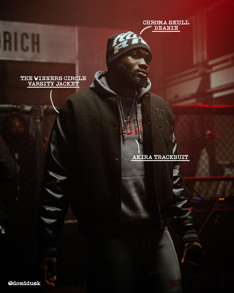 A man wearing a black varsity jacket with black sleeves, a gray hoodie, a black track suit, and a black beanie with a white and green graphic. White text labels point to different items: 'CHROMA SKULL VARSITY JACKET', 'AKIRA TRACKSUIT', and 'CHROMA SKULL BEANIE'.