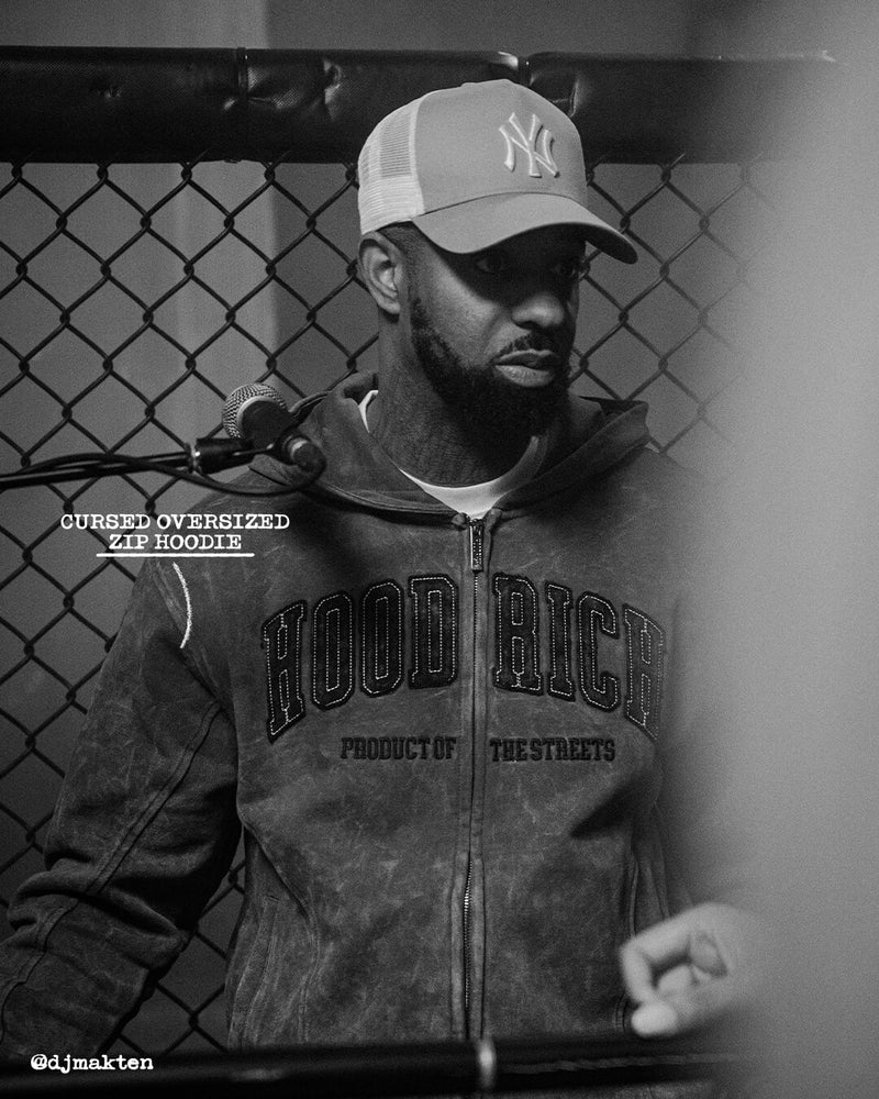 A black and white lifestyle photograph of a man wearing a distressed zip-up hoodie with 'HOODRICH' printed on the front, a New York Yankees baseball cap, and sunglasses, standing behind a chain-link fence.