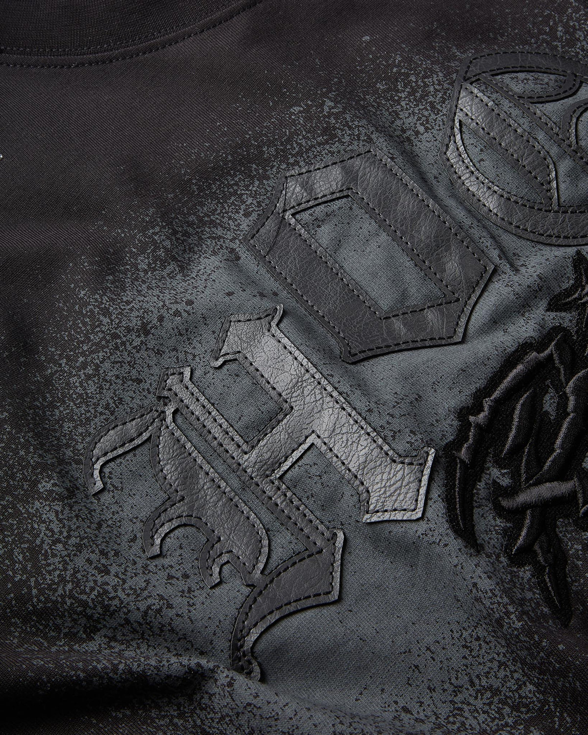 Close-up of a black textured garment featuring a large gothic-style embroidered letter 'H' with distressed, leather-like applique and tonal stitching.