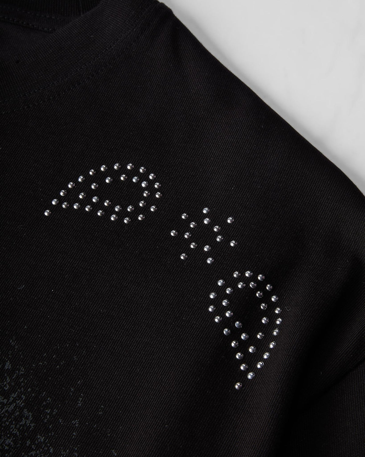 Close-up of a black knit garment with a rhinestone-studded logo on the chest, photographed against a light neutral background.