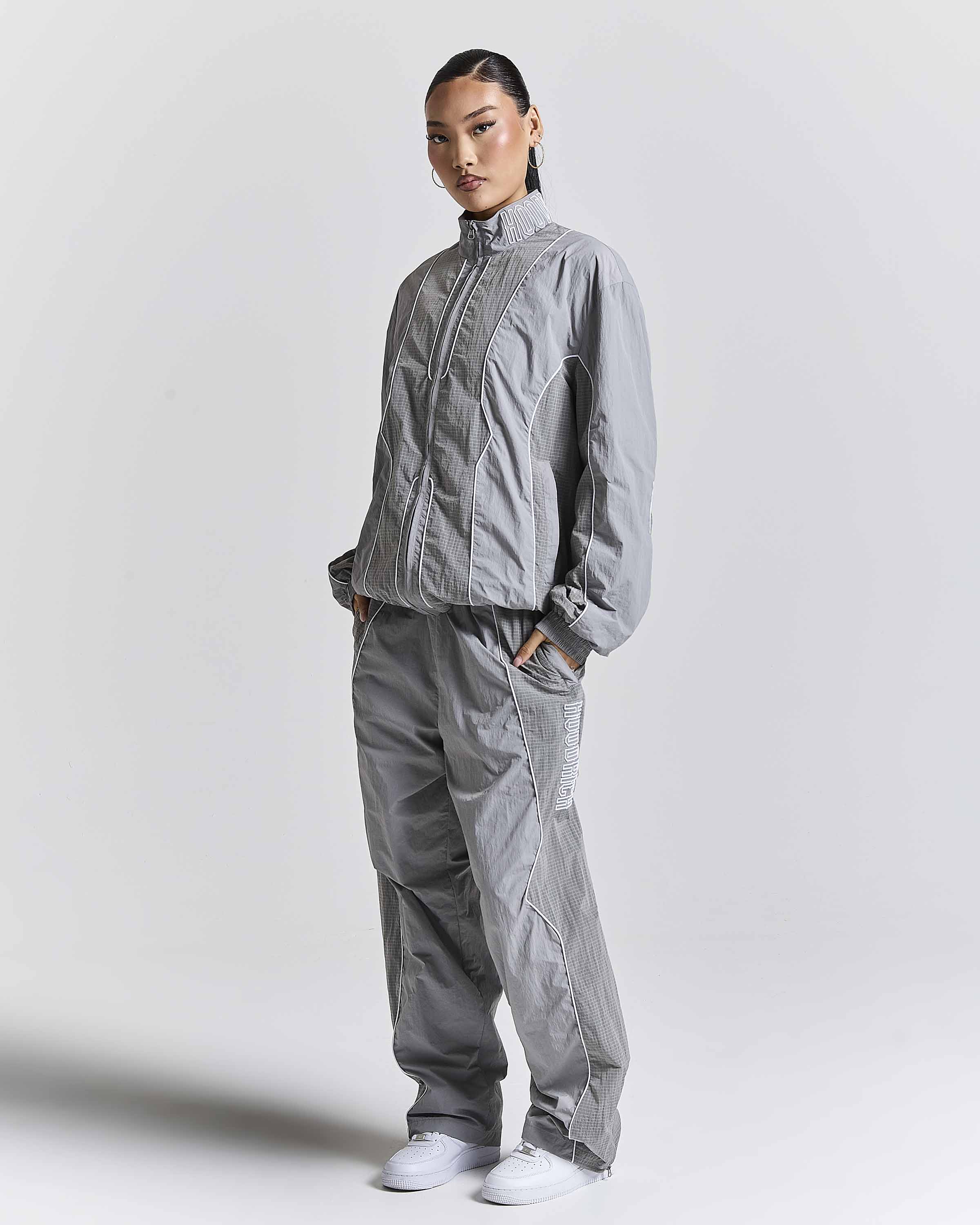 A model wearing a matching gray lightweight zip-up jacket and wide-leg pants set, styled with white sneakers, against a plain light gray studio background.