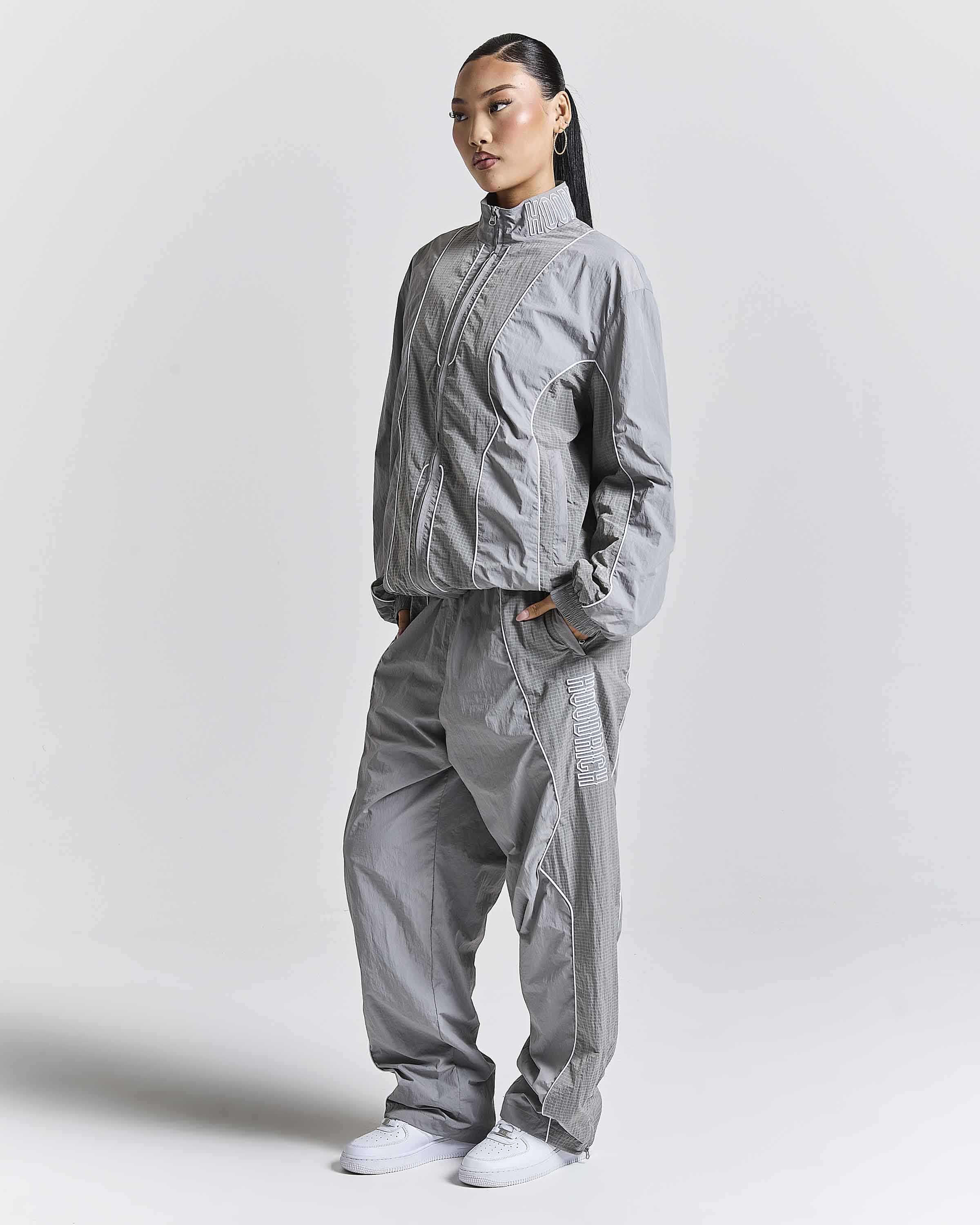 A model wearing a light gray technical windbreaker jacket and matching loose-fit pants, standing against a plain light gray studio background.