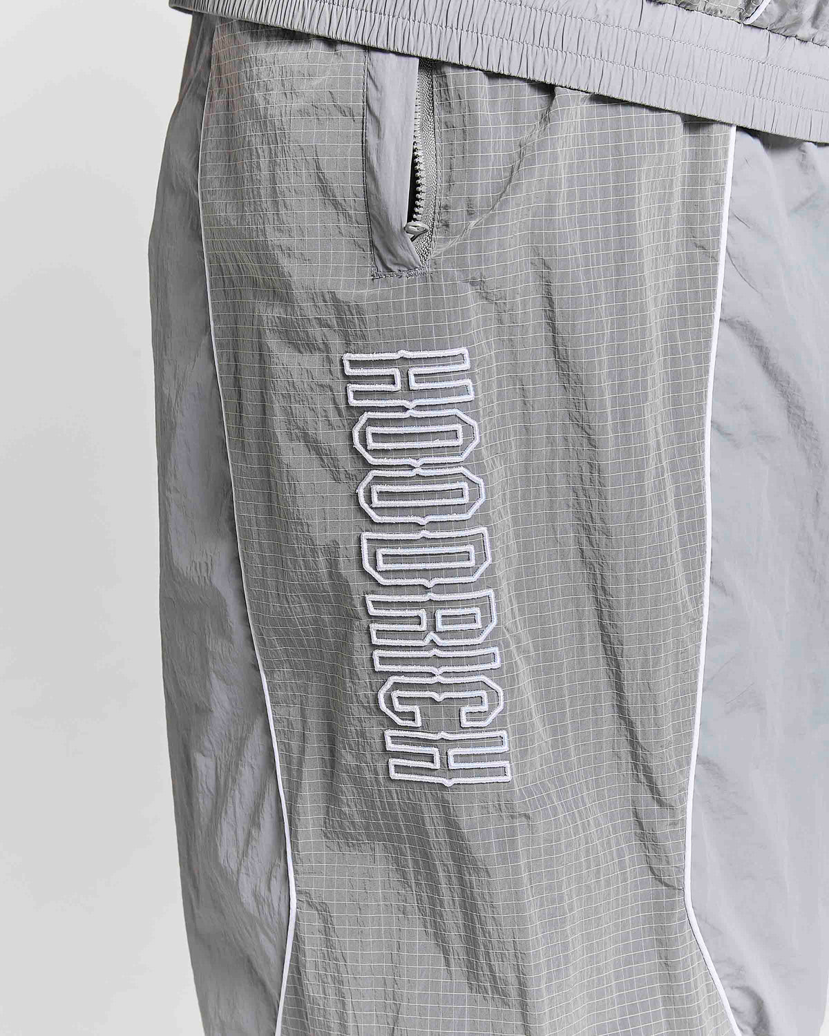 Close-up of gray HOODRICH windbreaker shorts with a white outlined vertical logo on the leg, featuring a zippered pocket and white piping.