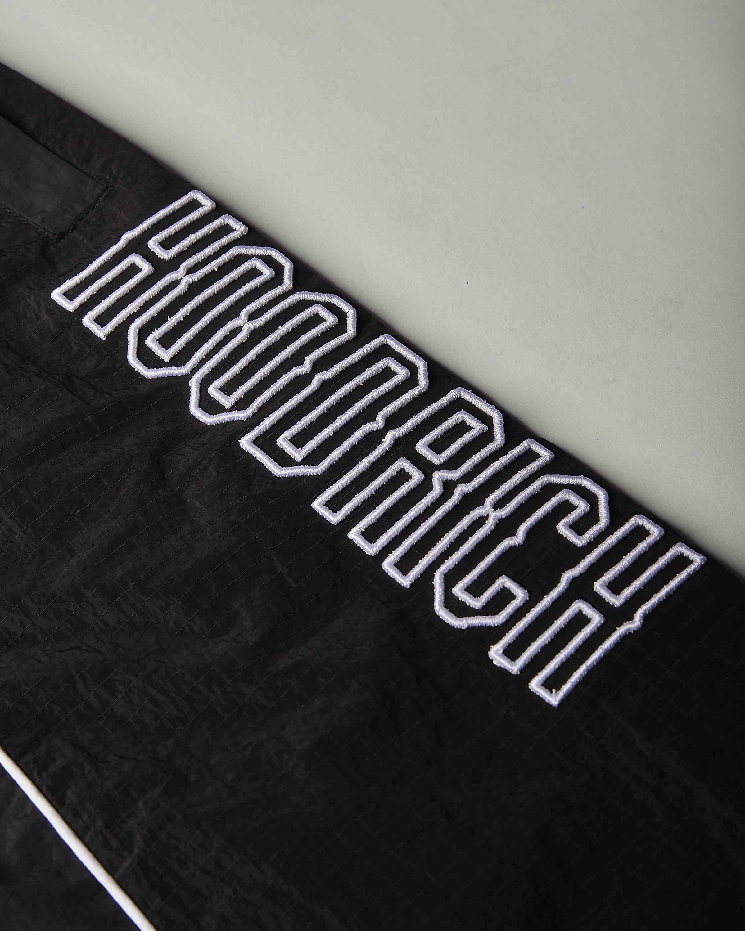 Close-up of a black textured fabric item featuring a large white embroidered 'HOODRICH' logo in a varsity-style outline font.